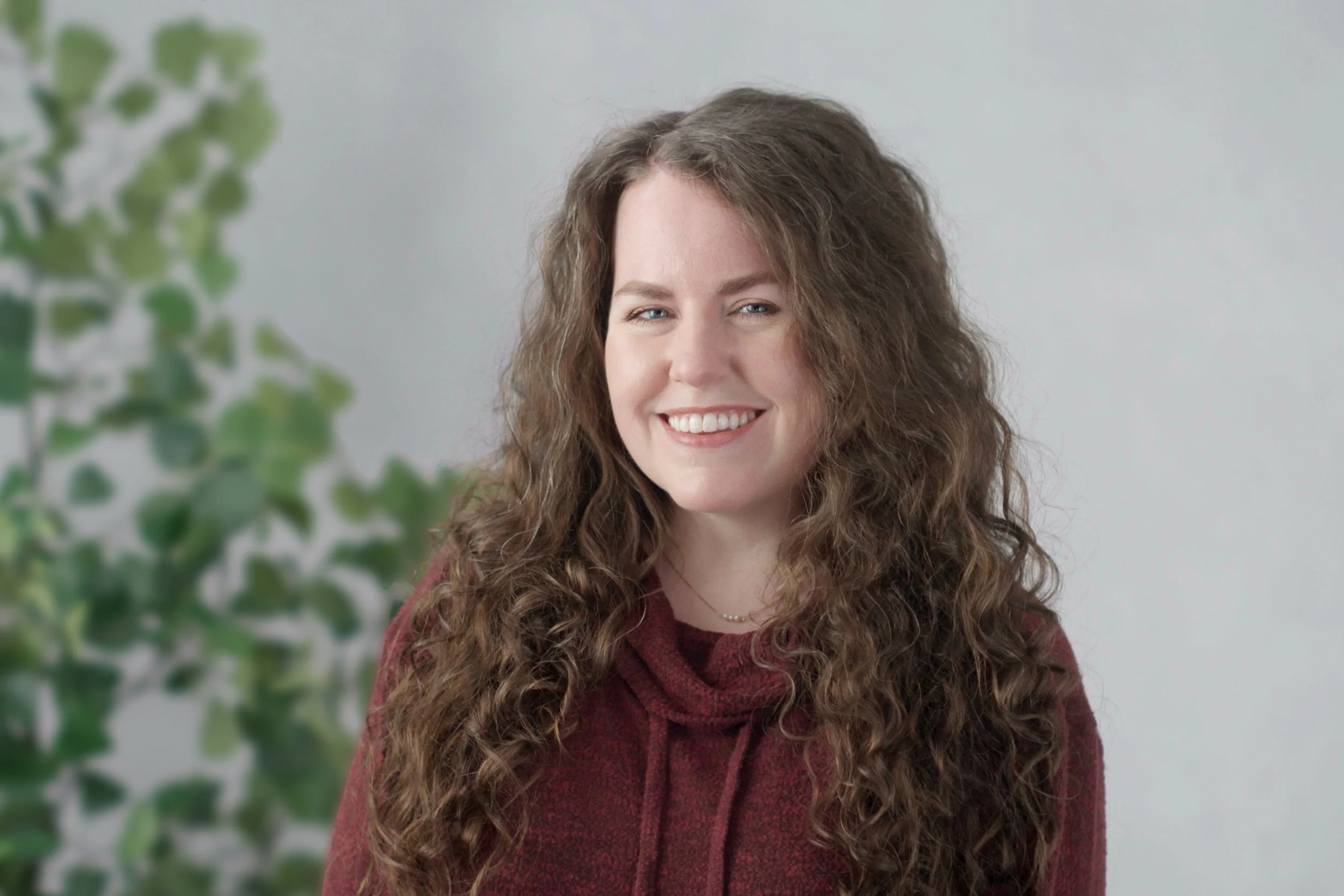 A woman with long curly brown hair, smiling, wearing a maroon sweater, standing indoors with a leafy plant on the left and a plain white background.