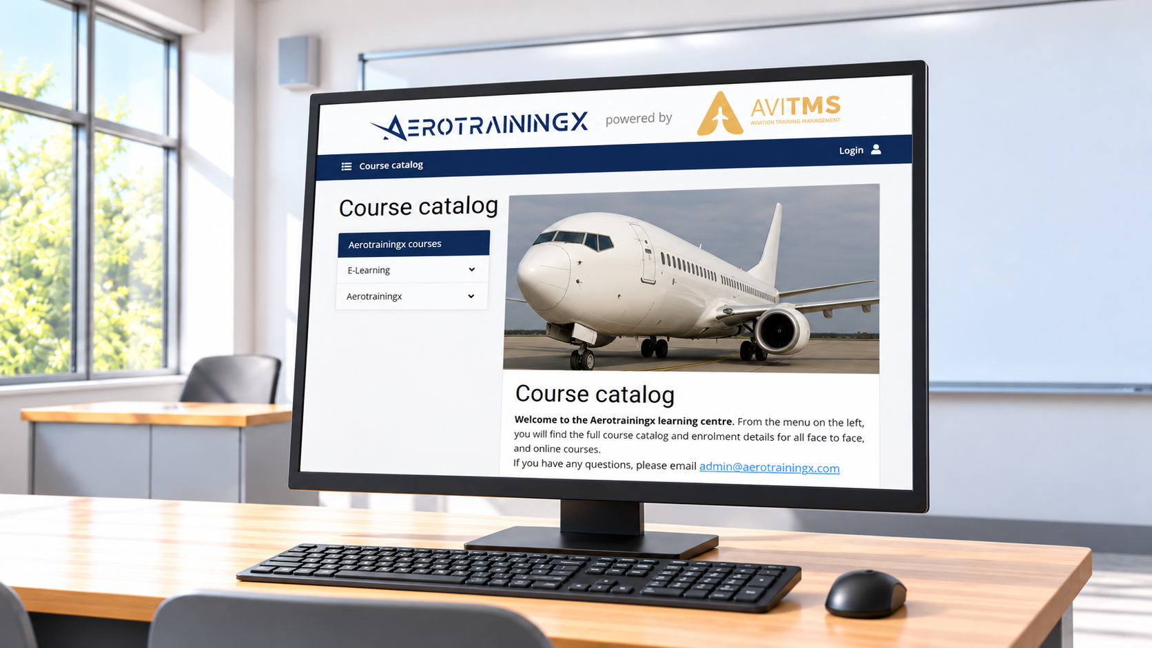 Computer monitor on desk displaying aerotrainingx course catalog with an airplane on the screen, in a conference room with large windows and a green outdoor view.