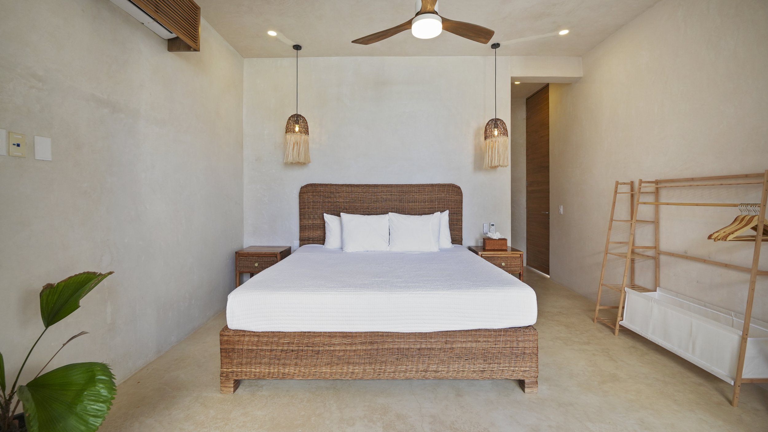 A bedroom with a large bed with white linens, rattan headboard, and two white pillows. There are two wicker bedside tables, colorful pendant lights hanging above, and a ceiling fan. The room has a beige wall, a wooden door, a plant in the corner, and a wooden clothing rack on the right side.