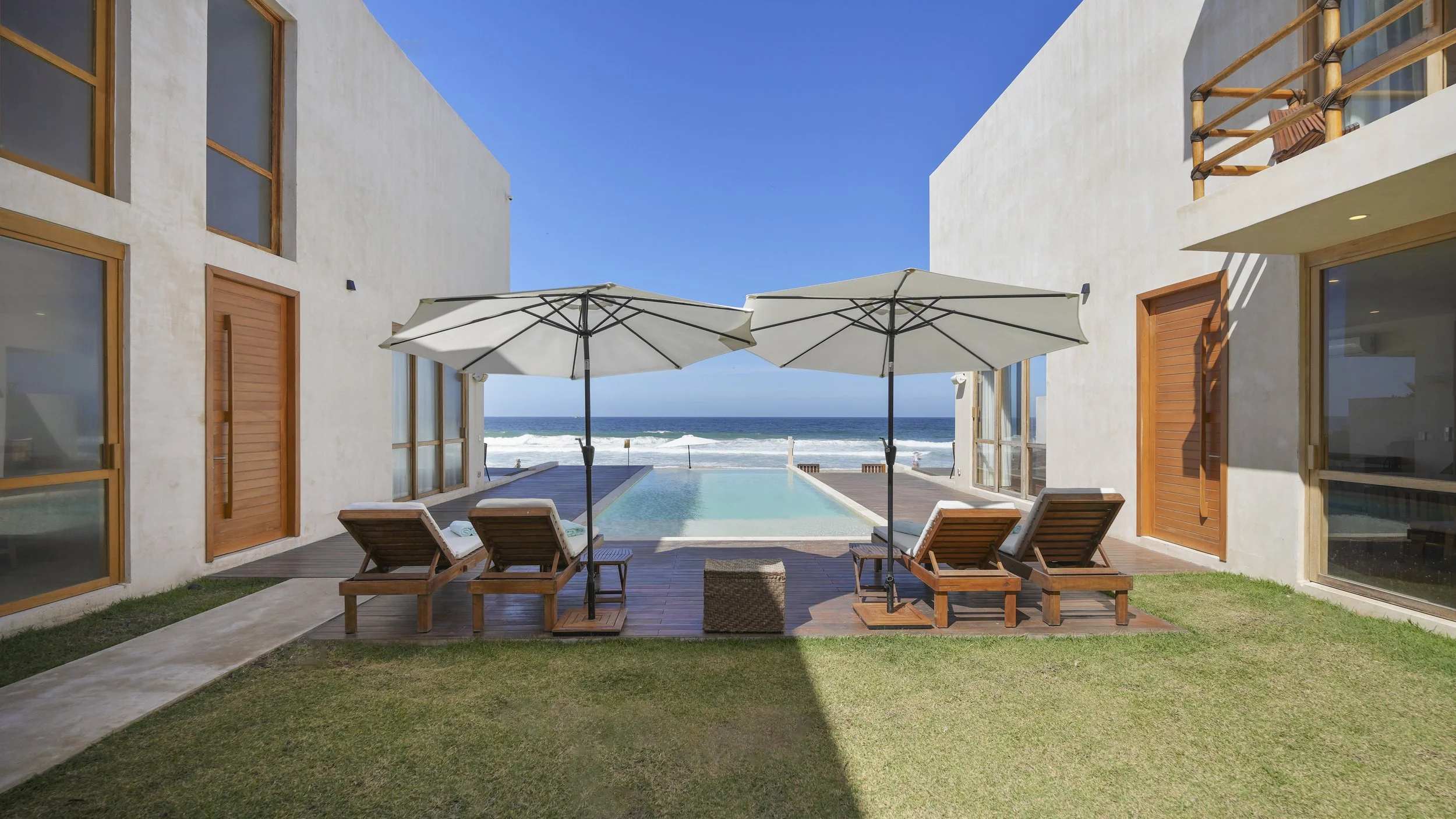 Luxury beachfront resort with swimming pool, two wooden lounge chairs with towels, two white umbrellas, ocean view, blue sky, modern white buildings with wooden accents.
