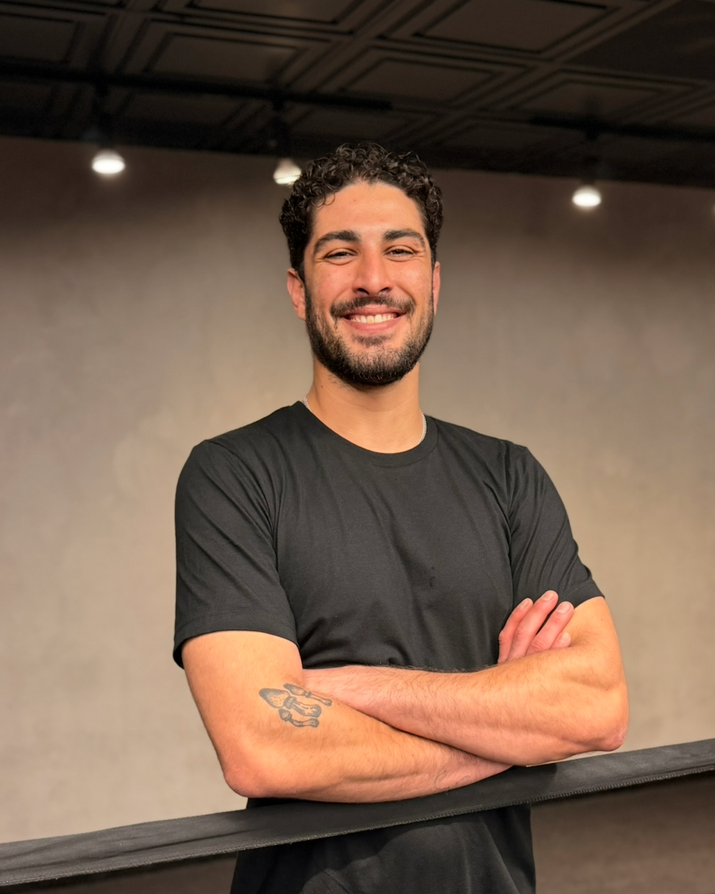 Smiling man with curly dark hair and beard, wearing a black t-shirt with crossed arms, standing indoors in front of a plain wall.