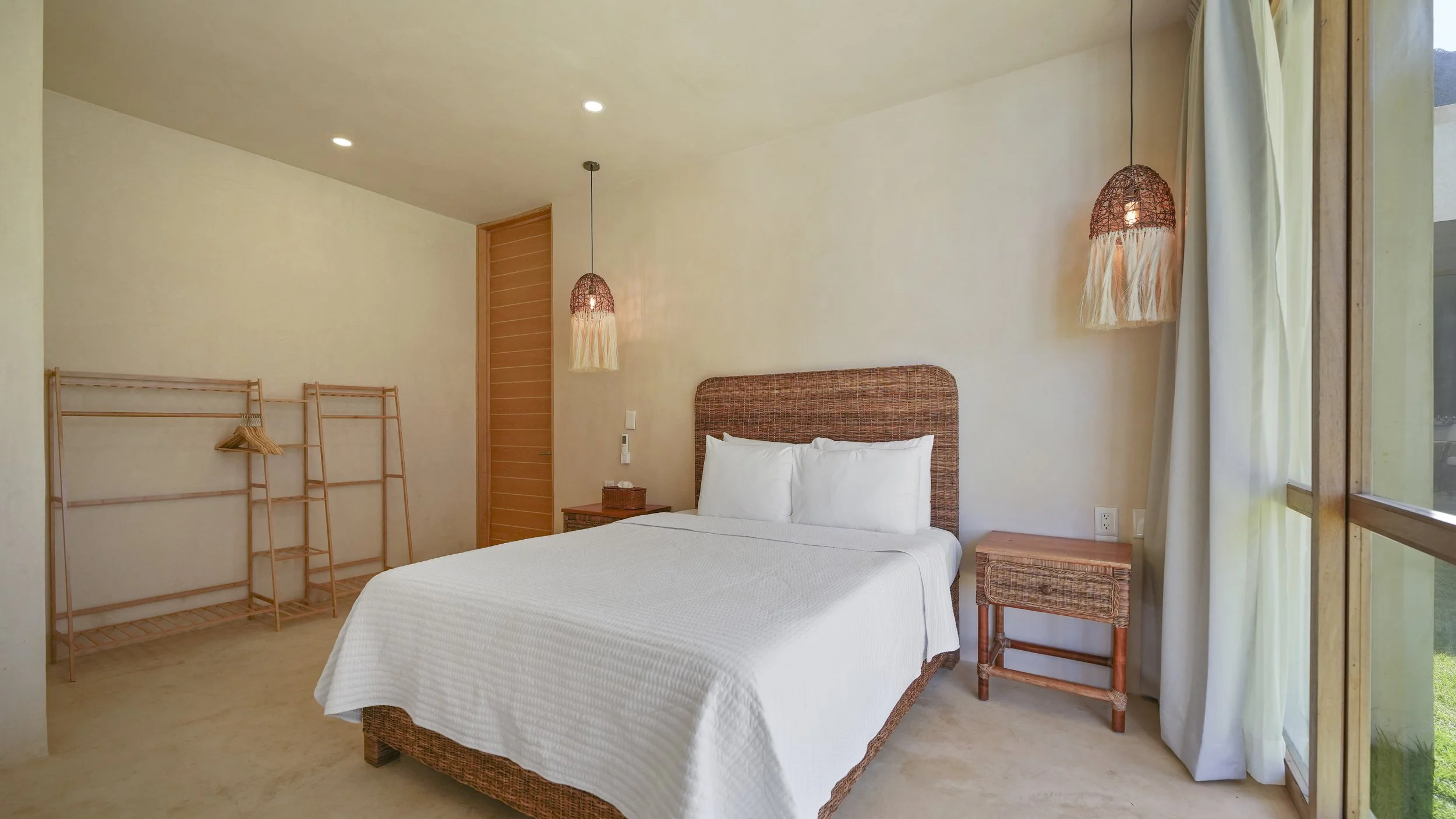 A modern bedroom with a bed featuring a rattan headboard, white bedding, and two side tables. There are two hanging lamps with woven shades and tassels, a closet with a wooden door, and a large window with curtains showing greenery outside.