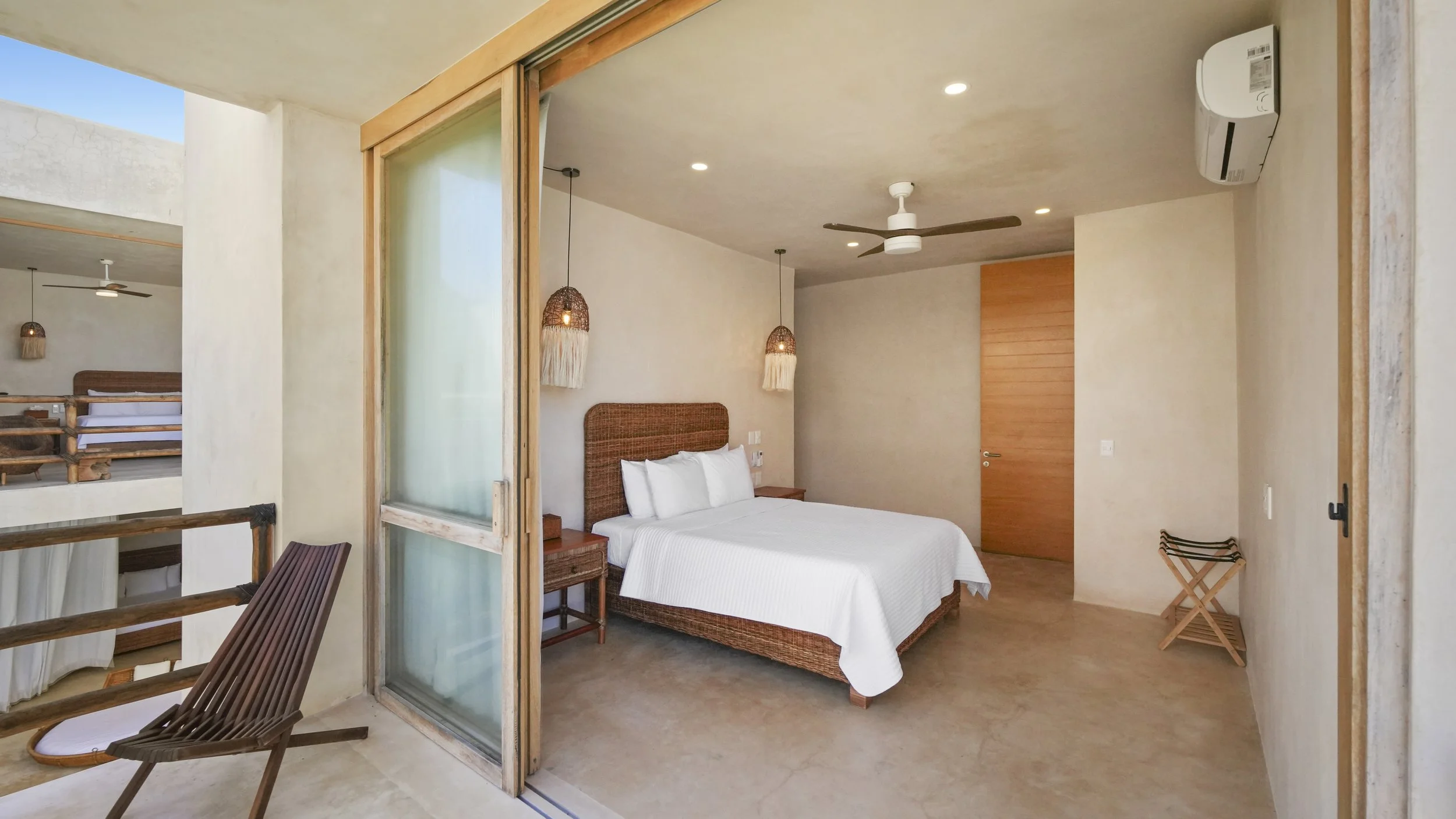 A bedroom with a large bed, wooden headboard, white bedding, two pendant lights with woven shades, ceiling fan, air conditioning unit, and a wooden door.