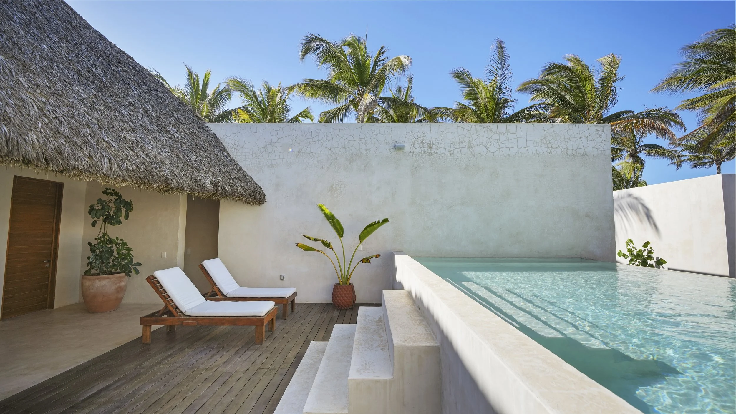 A private pool area with two lounge chairs on a wooden deck, tropical plants, and thatched roof structure, surrounded by white walls and palm trees under a blue sky.