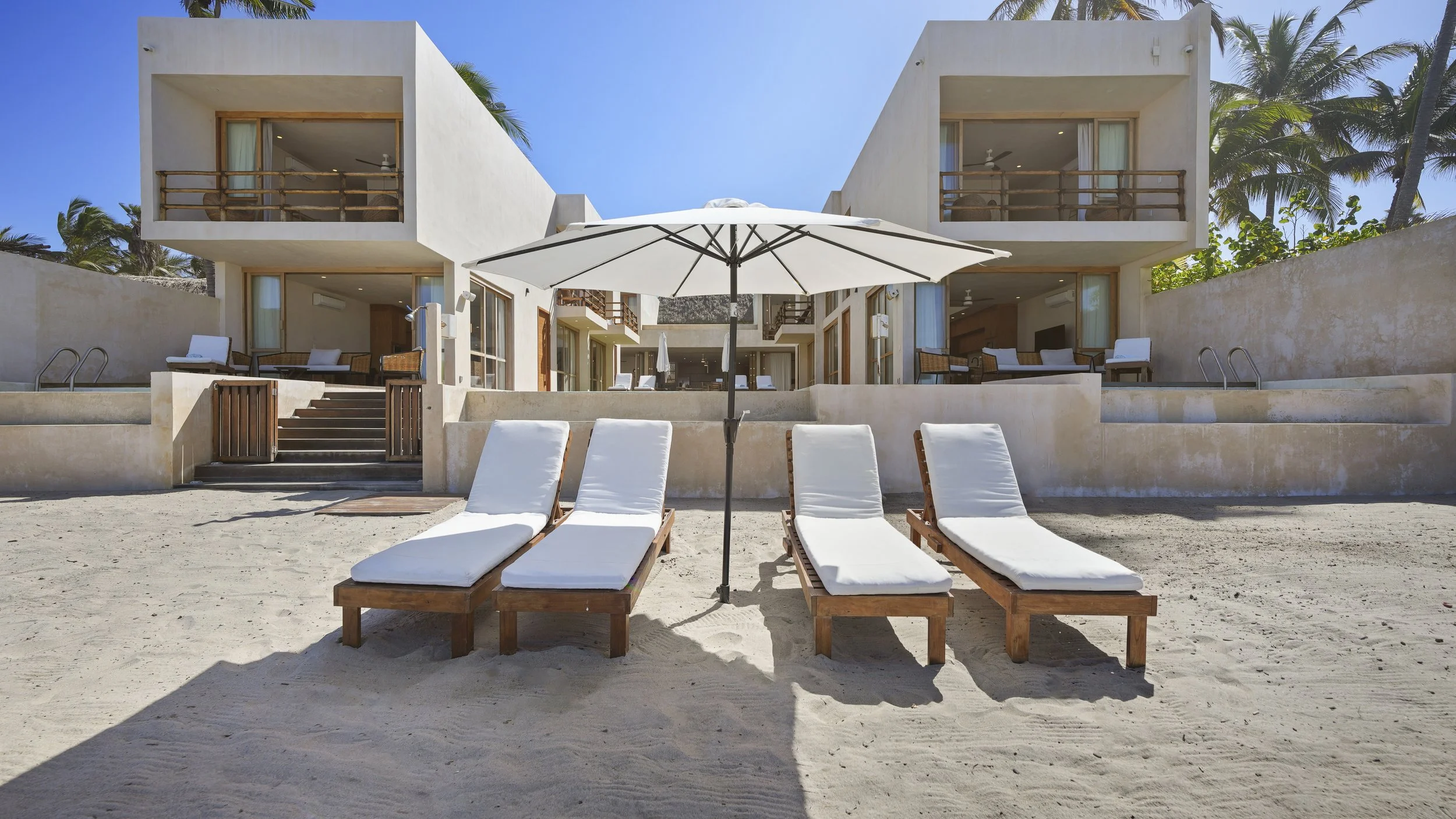 Beachfront resort with four white lounge chairs under a large white umbrella on sandy beach, modern white hotel building with balconies and outdoor seating in the background, palm trees, clear blue sky.