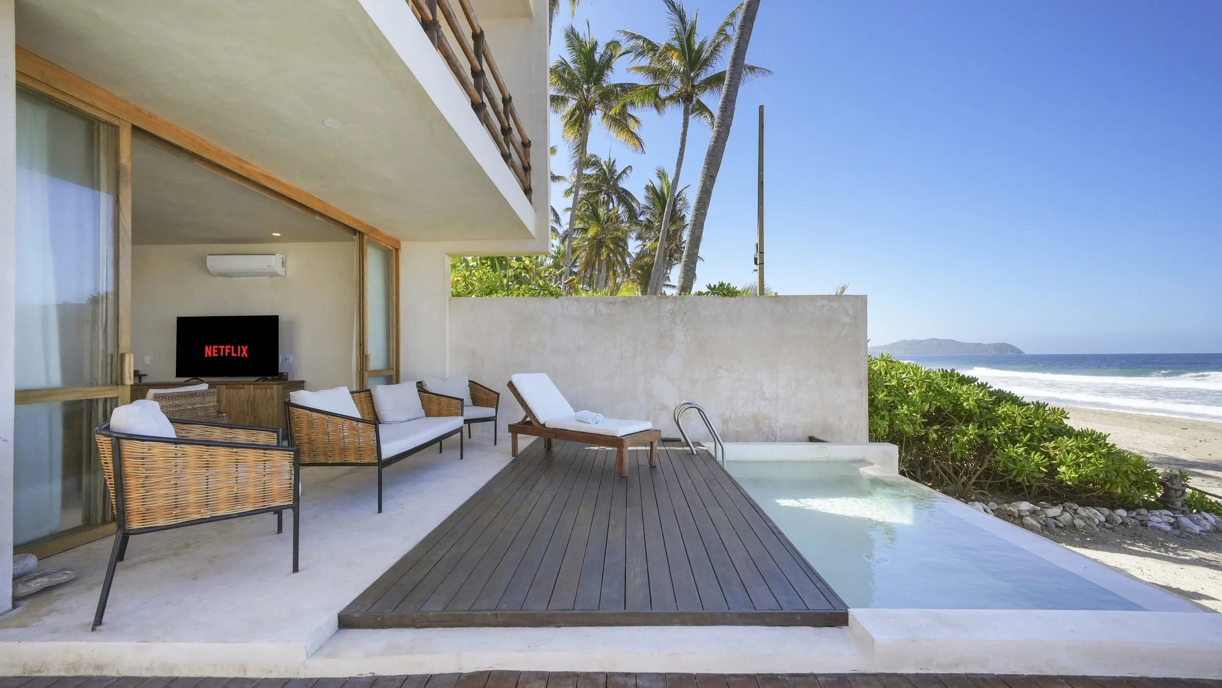 Outdoor balcony with couches, a lounge chair, and a small pool overlooking the beach with palm trees and ocean in the background.