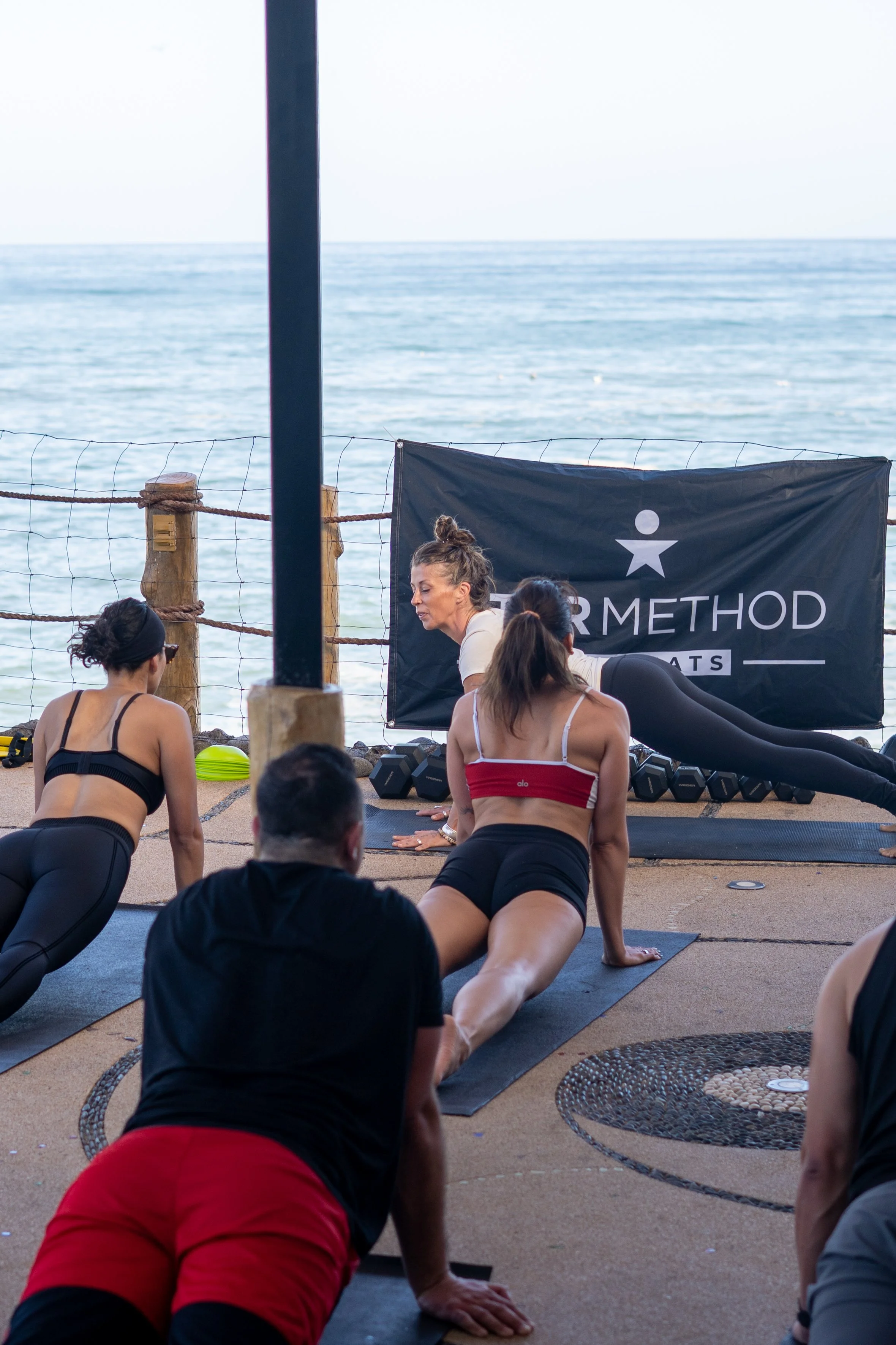 A group of people participating in a yoga class outdoors by the ocean, with an instructor leading the session and a black banner with 'PER METHOD' in the background.