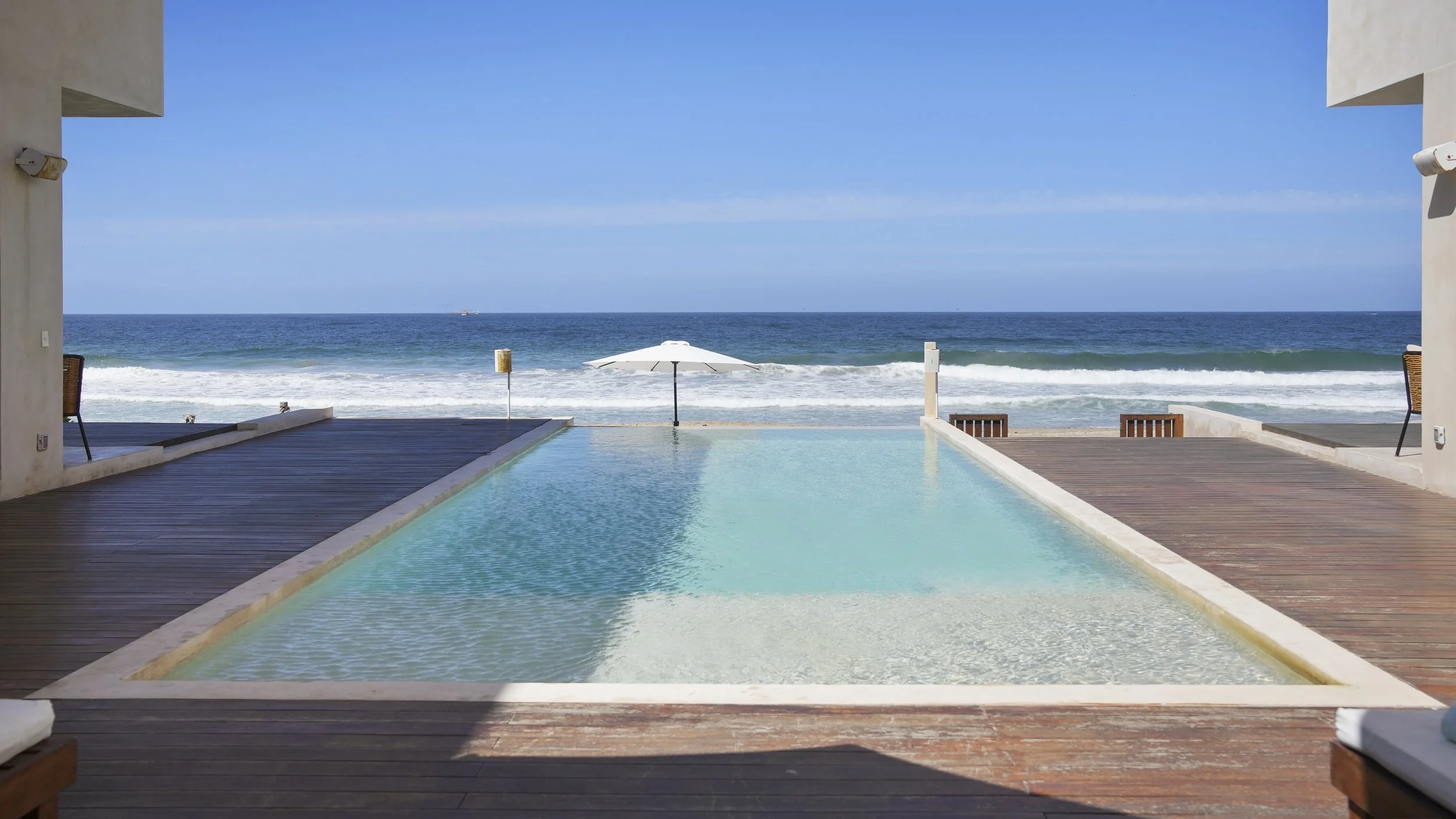 View of an infinity pool overlooking the ocean with waves, a white umbrella, and blue sky.