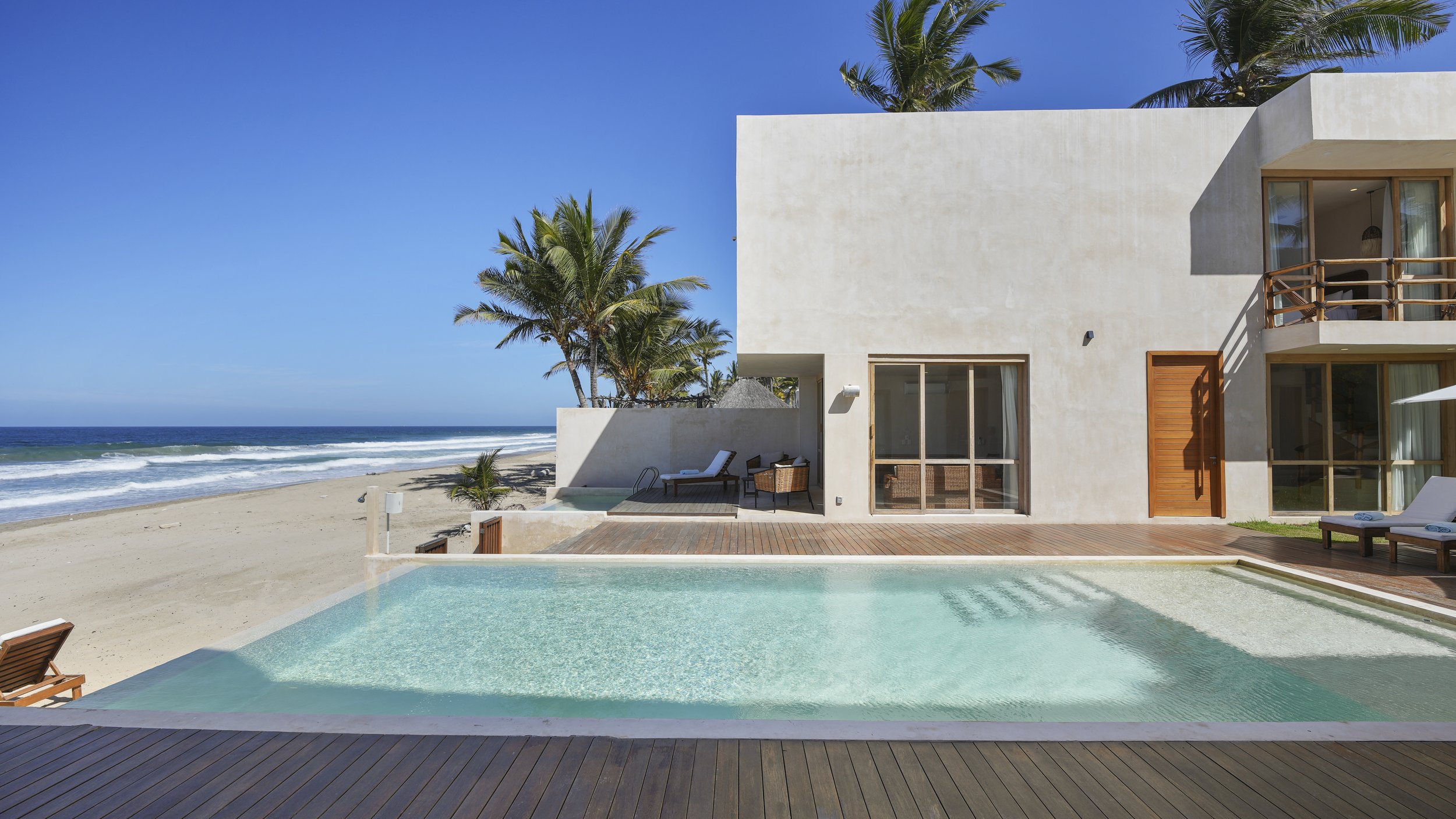 Luxury beachside house with swimming pool, deck chairs, palm trees, and ocean view under clear blue sky.