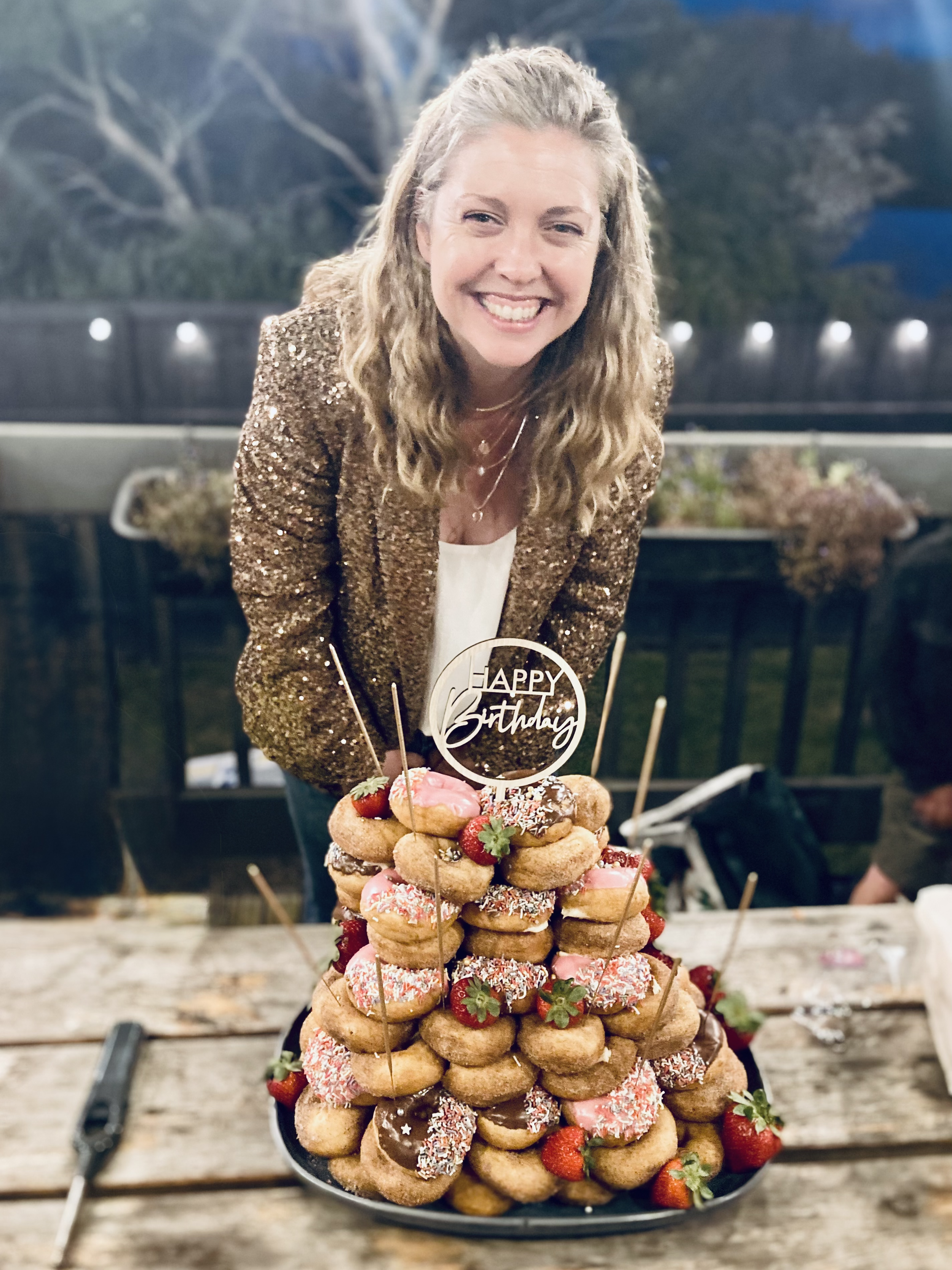 Woman in a gold sequin blazer smiling behind a large birthday doughnut tower with strawberries and pink and chocolate frosting, topped with a "Happy Birthday" sign.