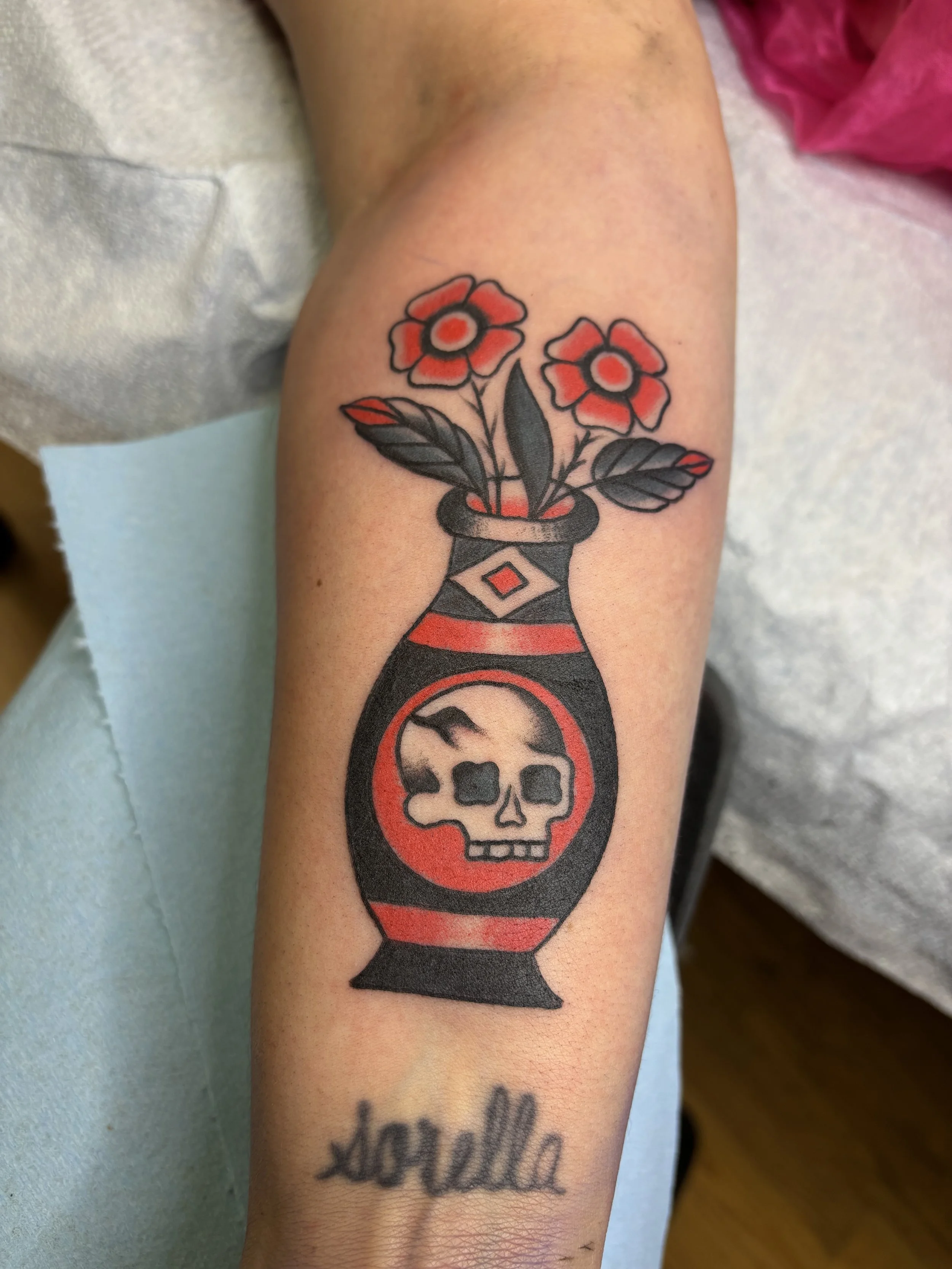 Tattoo of a black and red vase with a skull and mountain design, holding red flowers with black leaves, and the word "adorella" written below.