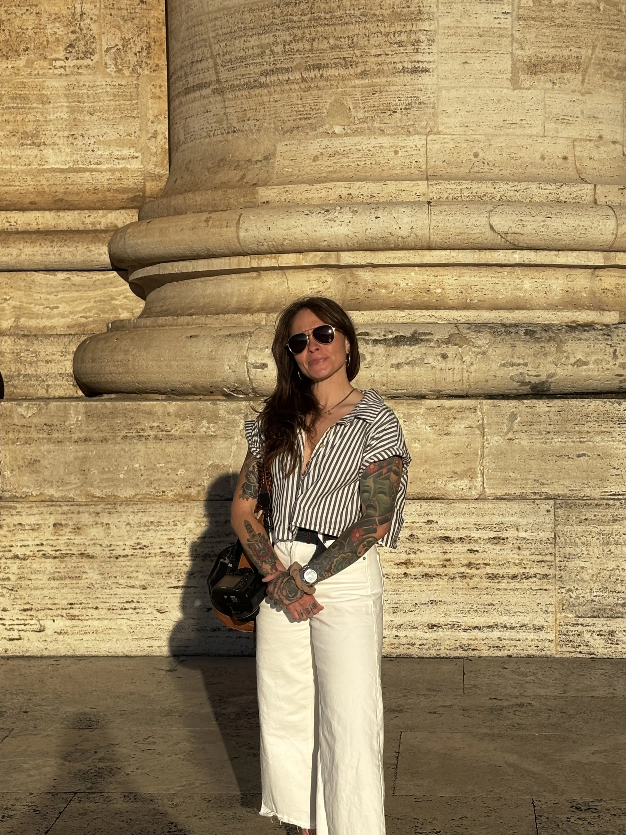 A woman with tattoos, wearing sunglasses, a striped shirt, and white pants, standing in front of a large stone column at sunset.