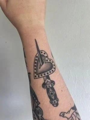 Tattoo of a heart-shaped locket with a needle passing through it on a person's arm.