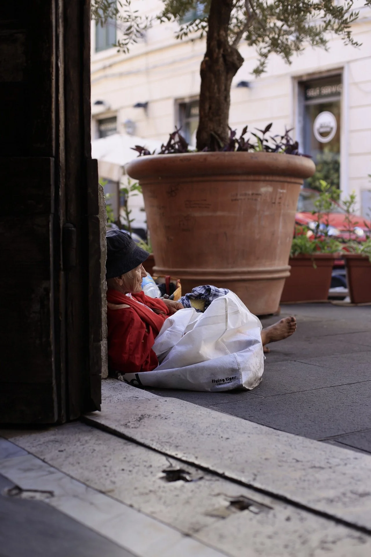 A person with a black hat, red jacket, and white bag sitting on the sidewalk leaning against a wall, with a large plant in a terra-cotta pot behind them.
