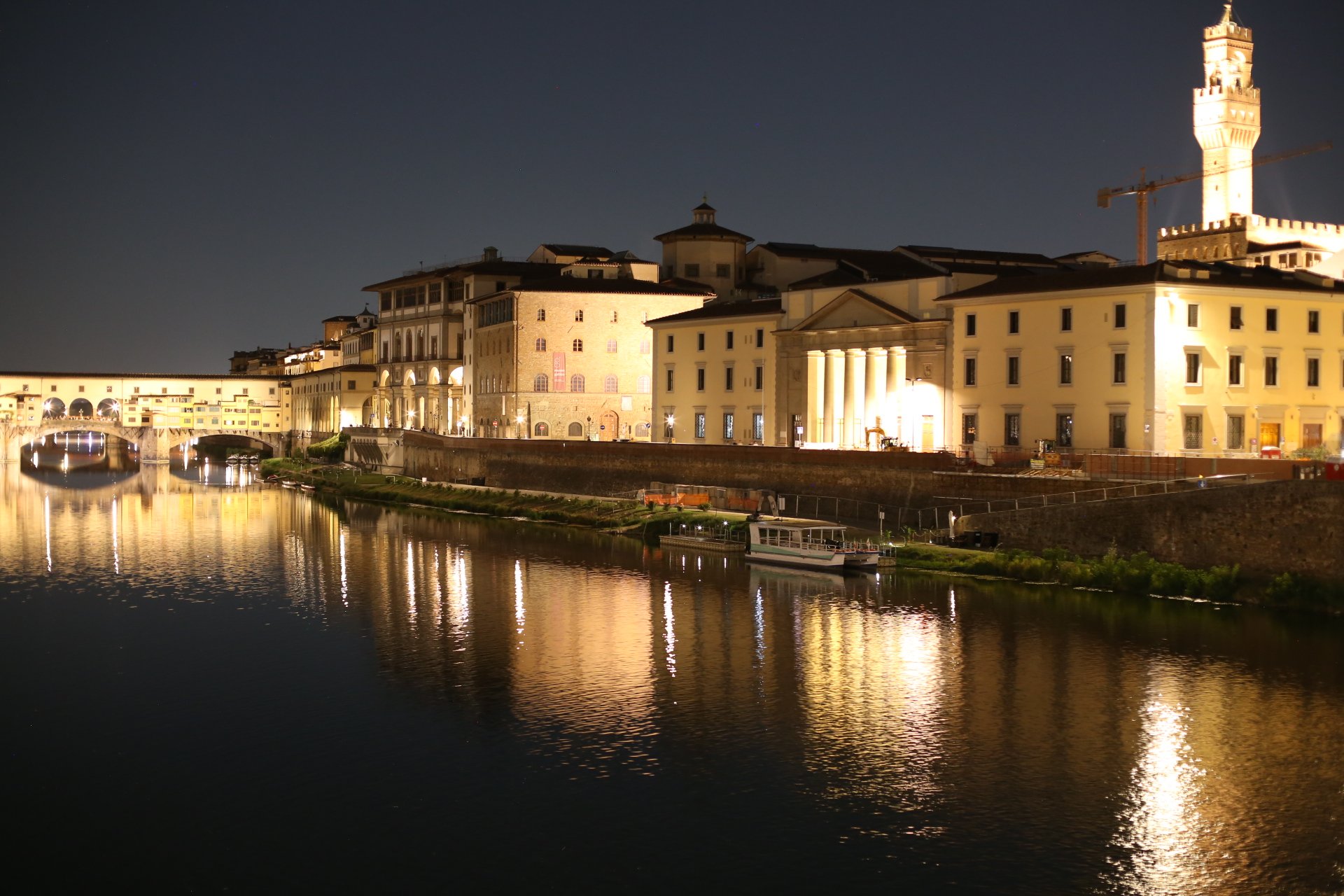 Night view of historic buildings along the river in Florence, Italy, with reflections on the water and illuminated structures, including a tall tower.