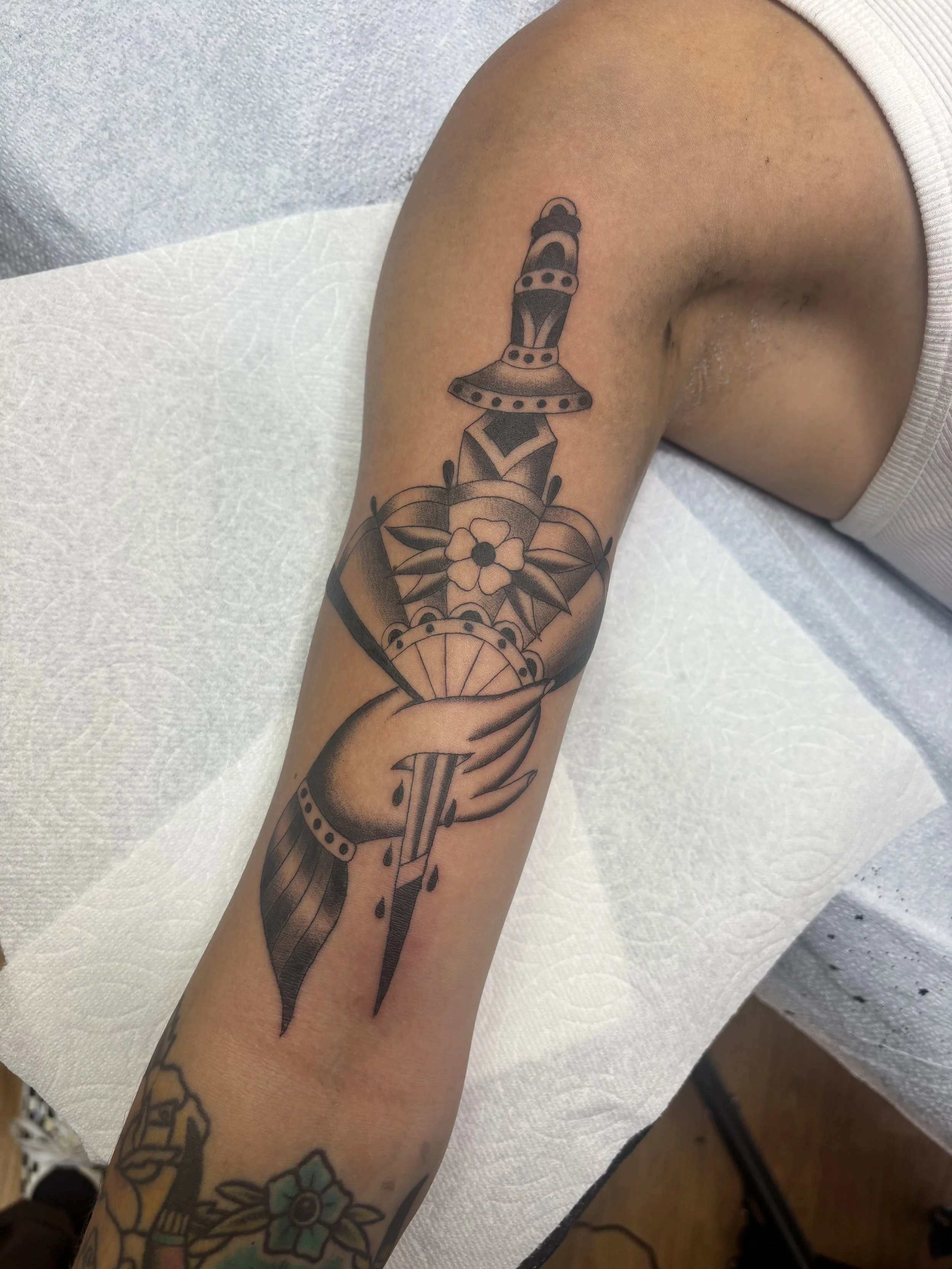 A black and gray tattoo of a dagger piercing a flower, with a hand holding the dagger, on a person's forearm.