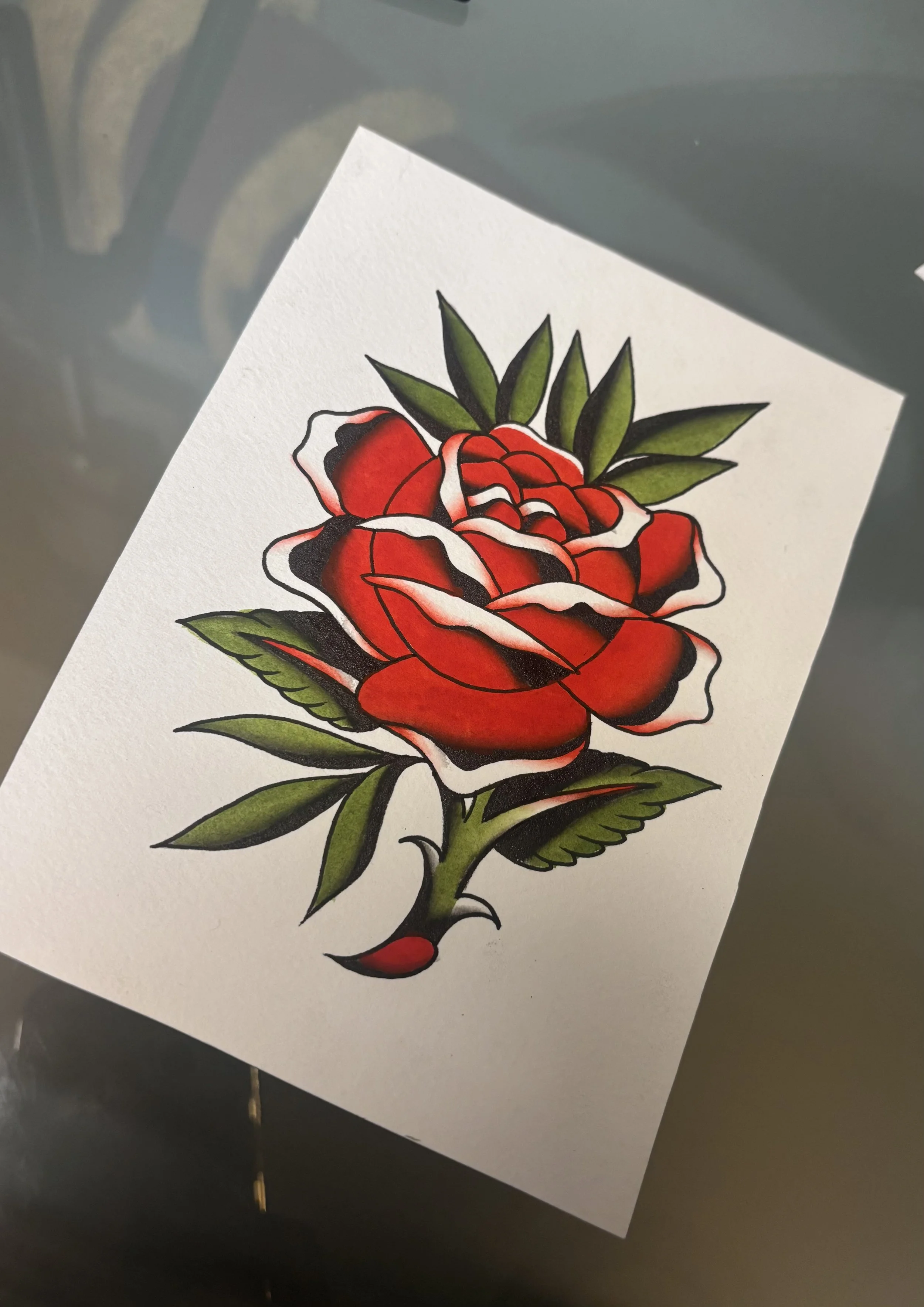 A tattoo design of a red rose with green leaves on white paper.