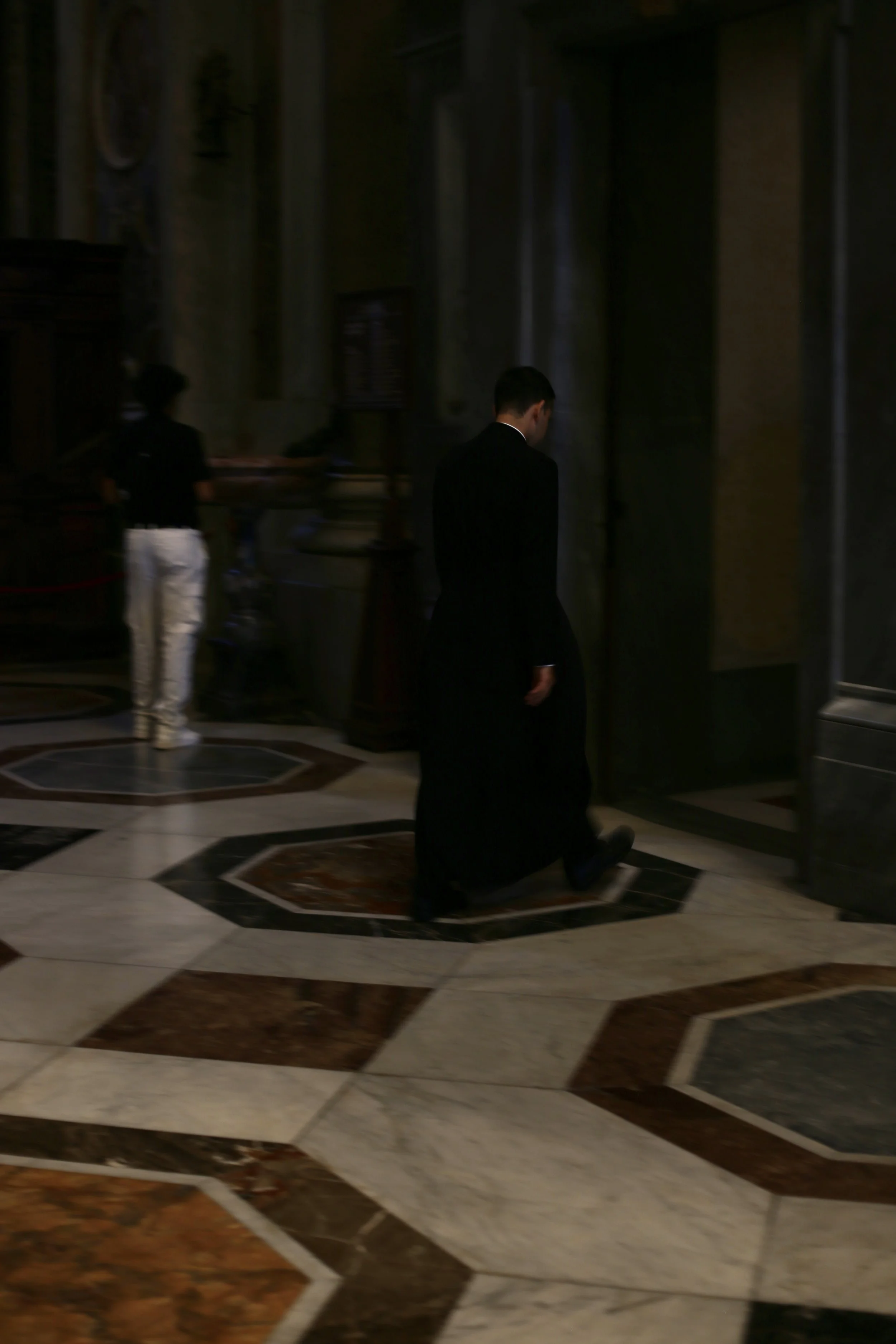 Man in a black suit walking in a dimly lit, ornate interior with patterned marble floors.