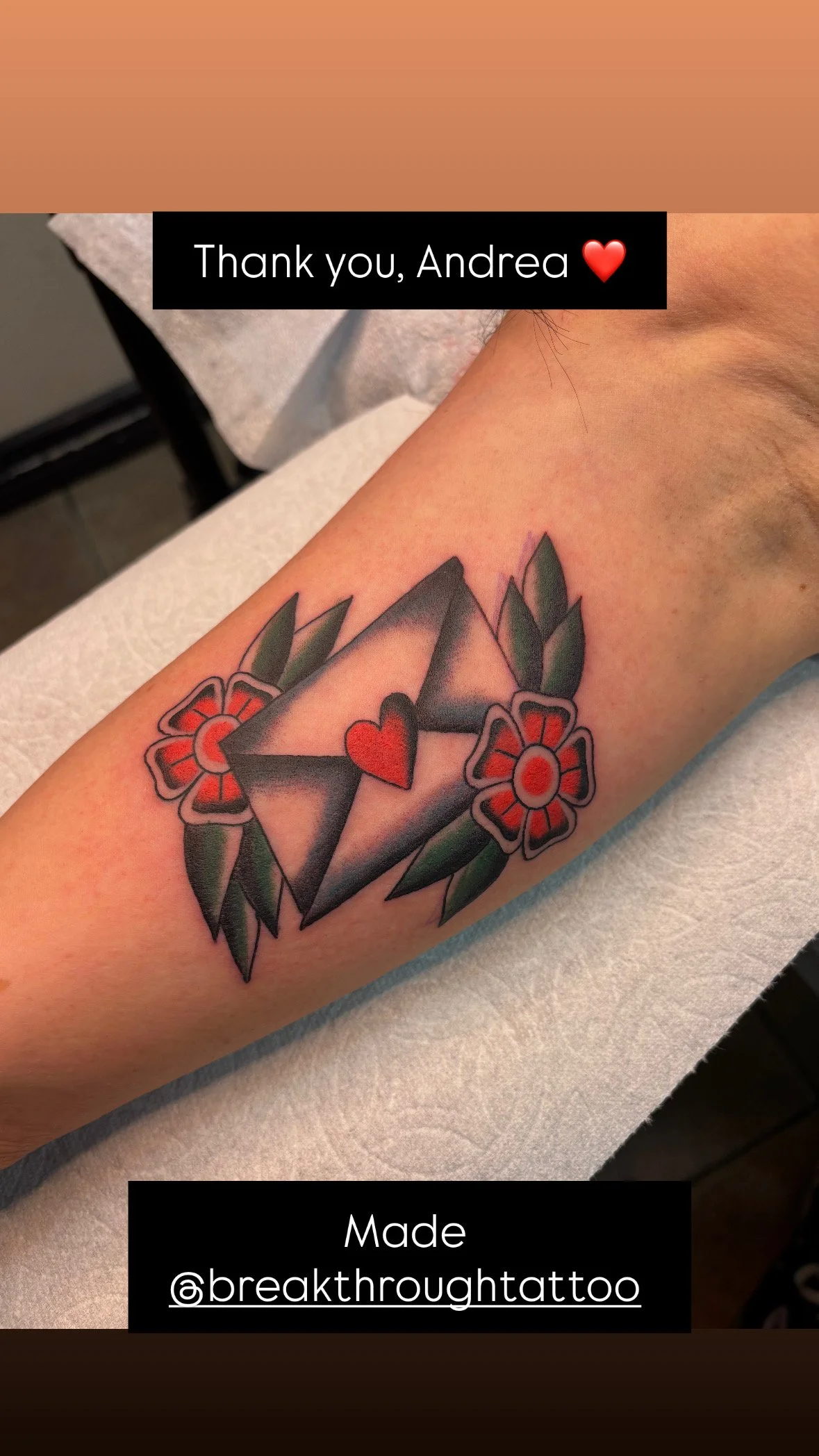 Tattoo of a geometric 3D diamond with a red heart inside, surrounded by two red flowers with green leaves.