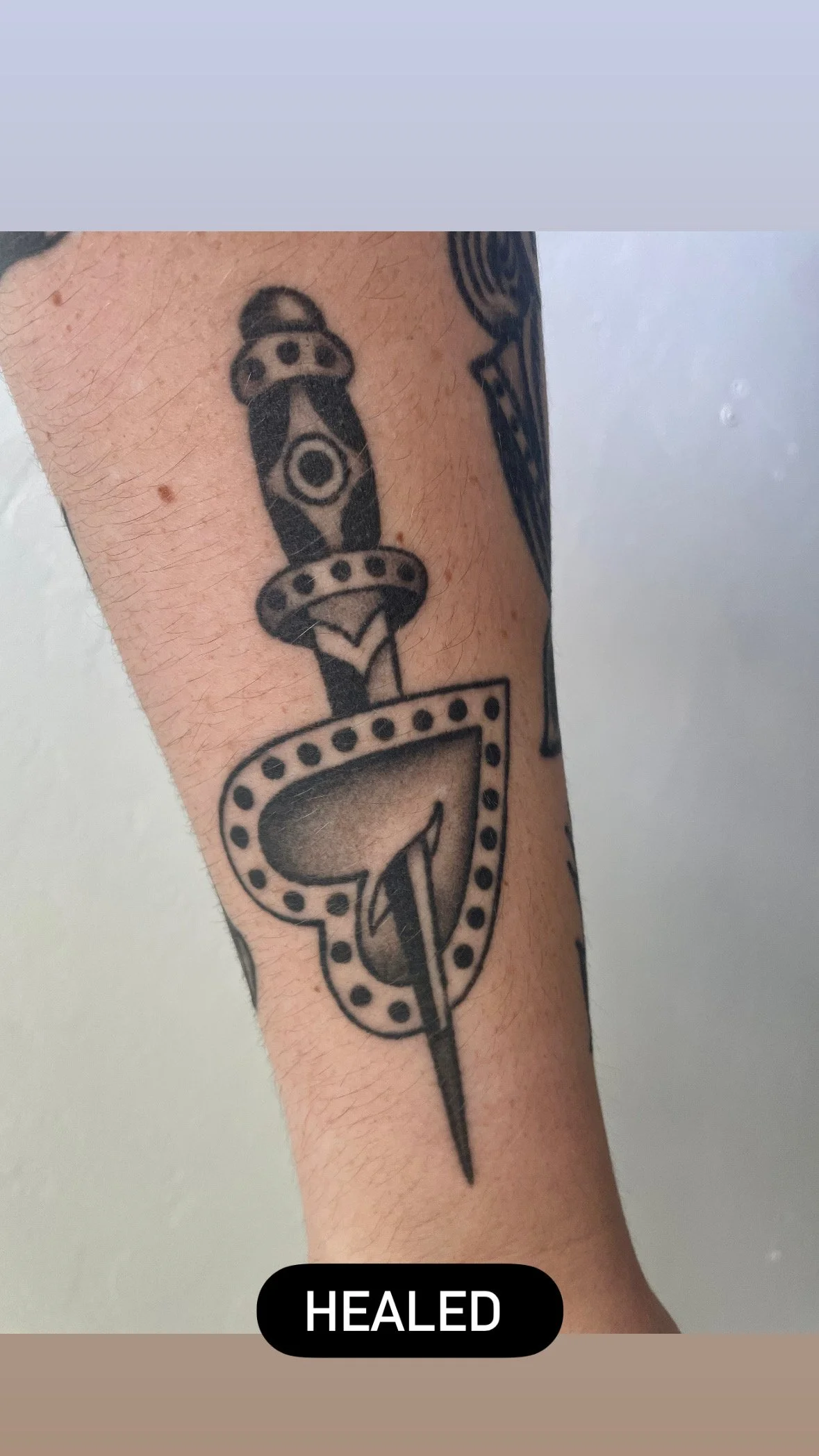 Tattoo of a stylized dagger with a heart-shaped top on a person's forearm, with the word 'HEALED' across the bottom.