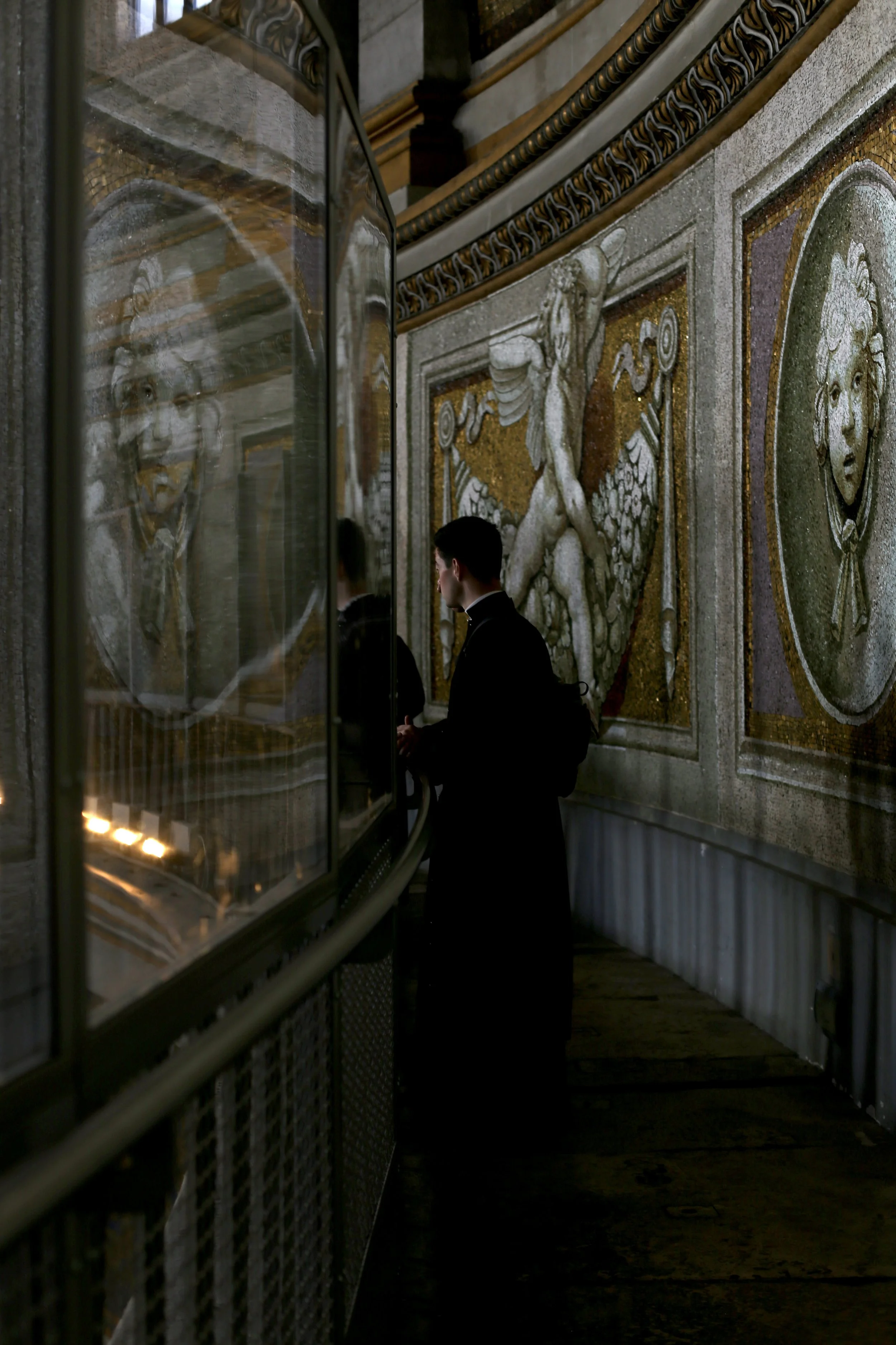 A man dressed in black standing in front of a glass display case inside a building with ornate, mosaic wall decorations including a griffin and a lion.
