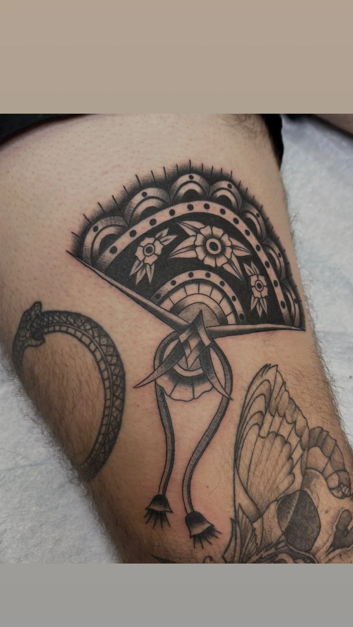 Tattoo of a decorative fan, a snake, a butterfly, and part of a flower on a person's thigh.