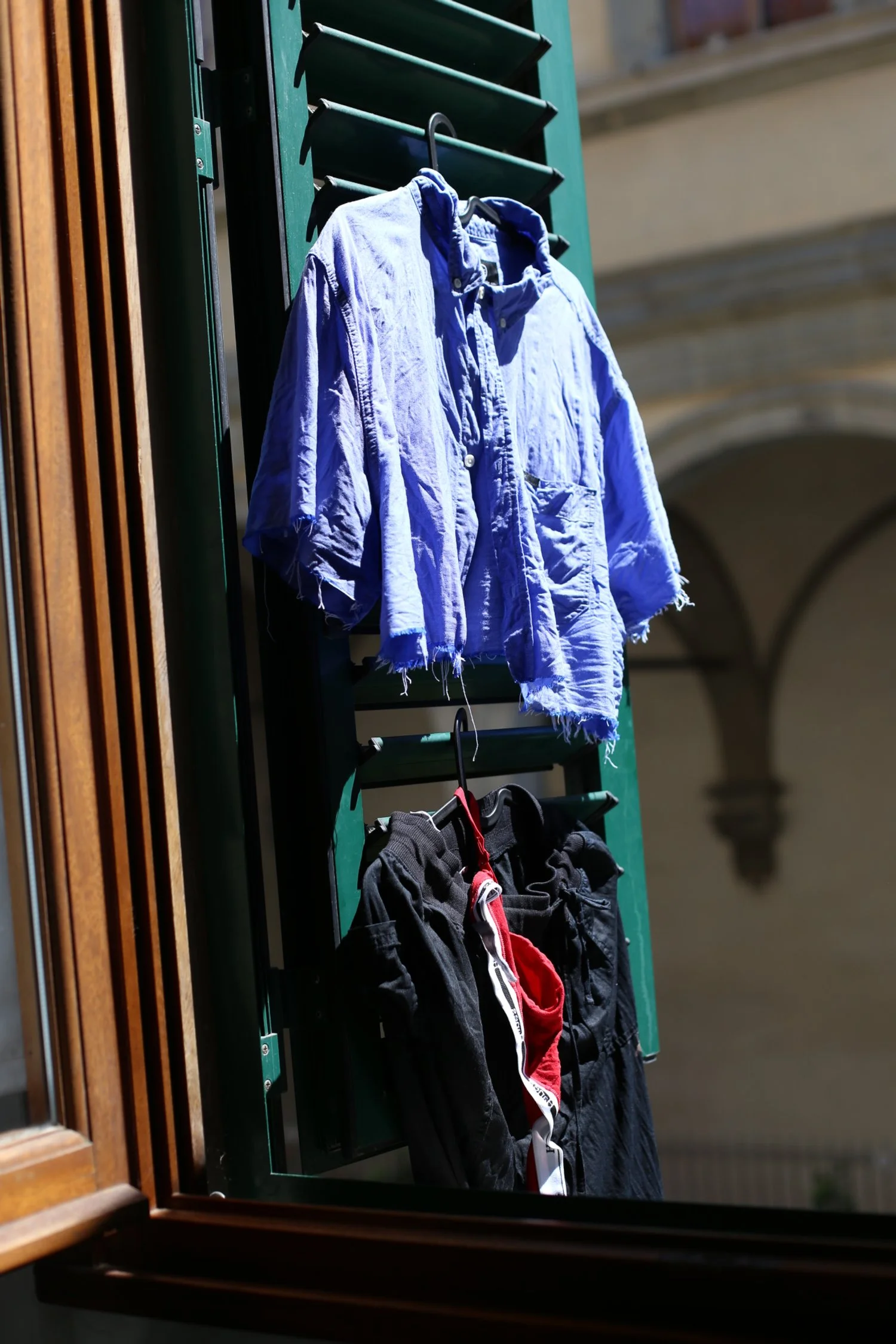 A blue, frayed, long-sleeved shirt hanging outside on a black hanger, attached to a green metal structure, with a pair of black pants and red-and-white towel hanging below.