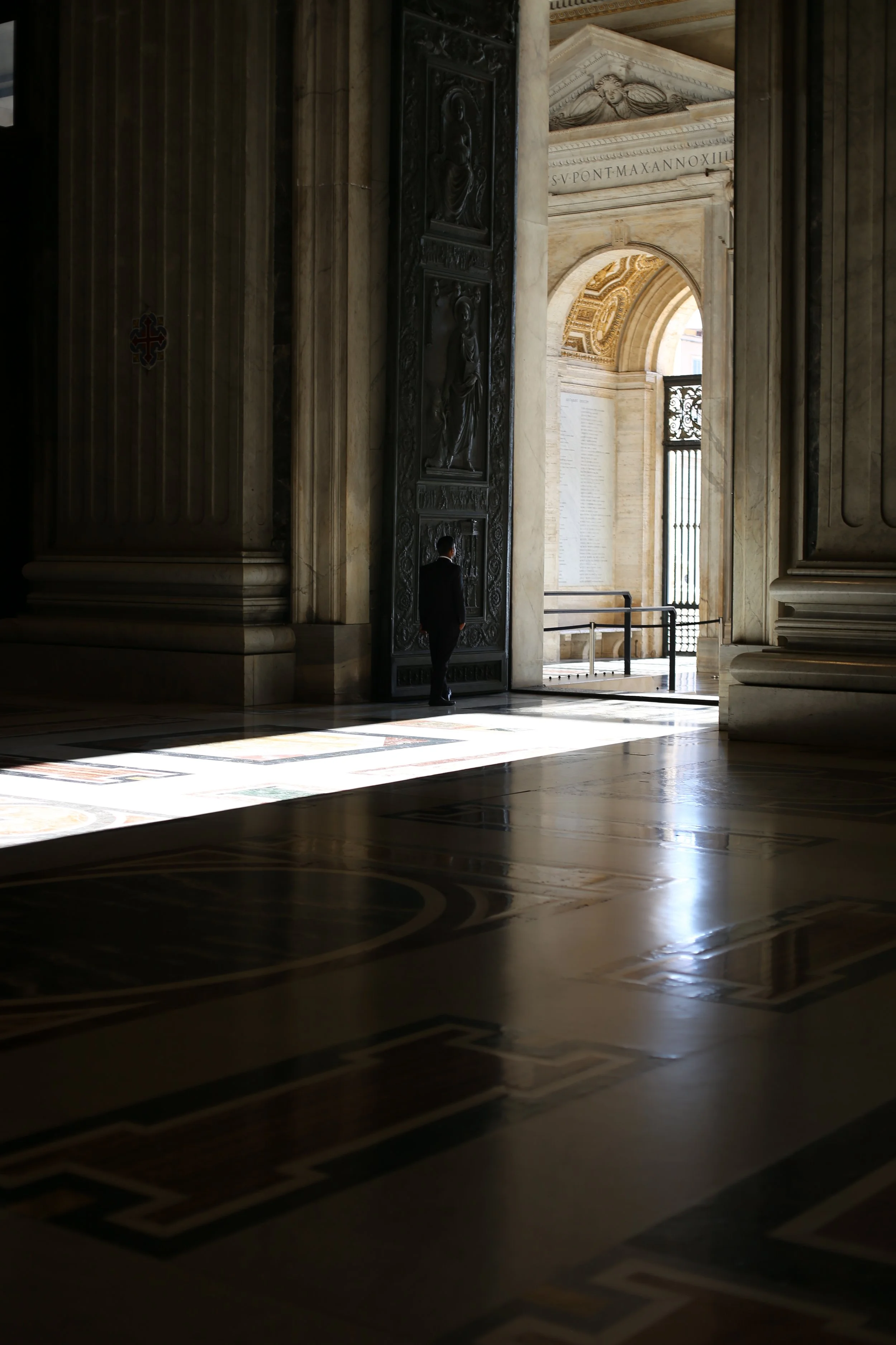 Interior of a grand building with classical architecture, light streaming in through doorways, a man standing near a large decorated door, and intricate patterned flooring.
