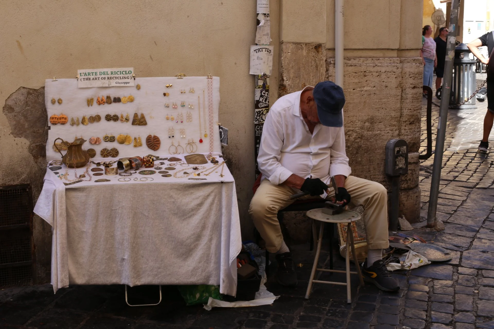 Street vendor sitting on a stool, working with jewelry or small metal items, with a table displaying jewelry and accessories, against a tan wall and cobblestone street.