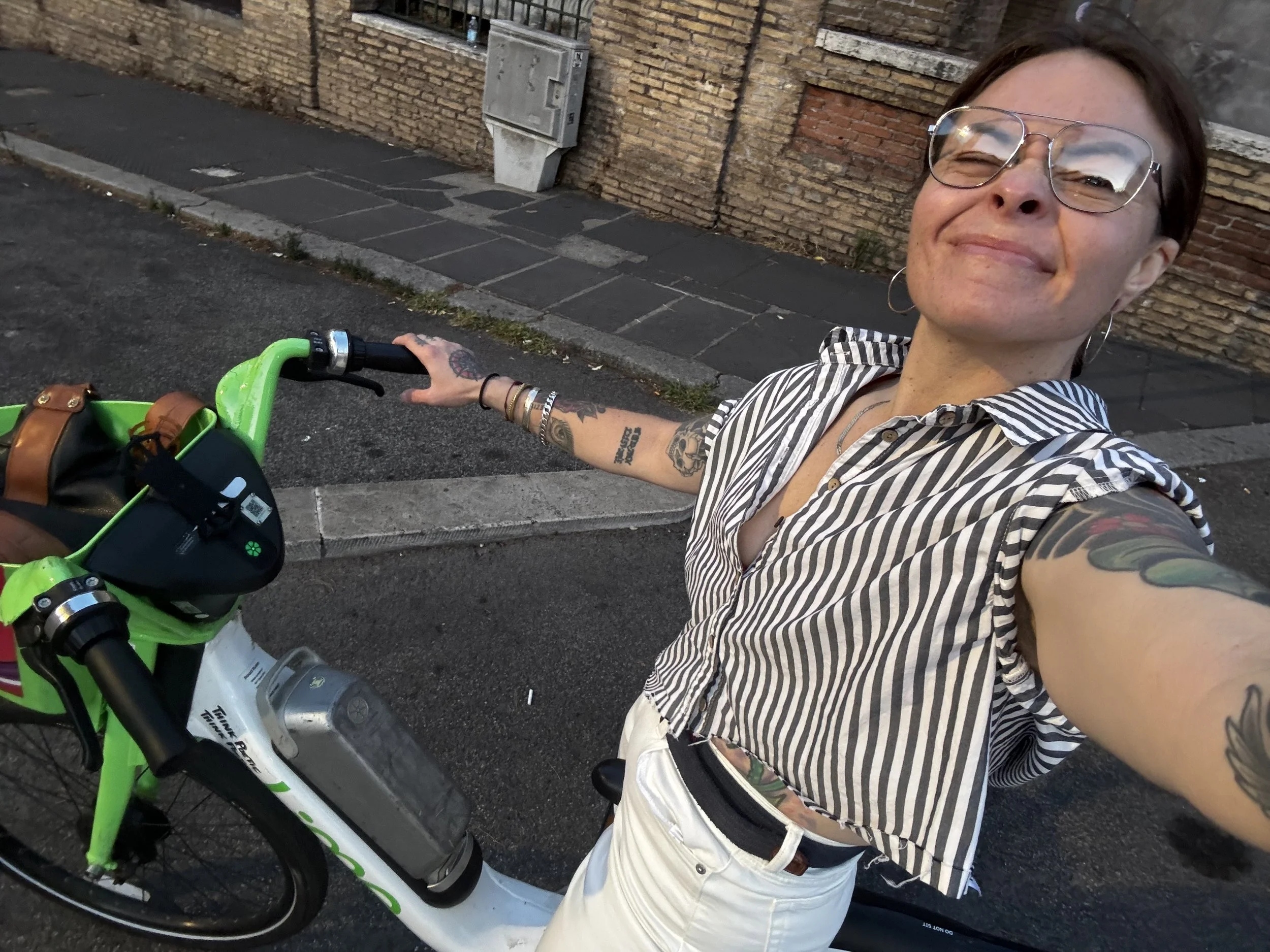 A woman with short brown hair, glasses, and tattoos on her arms takes a selfie with her electric bike on a city street, smiling.