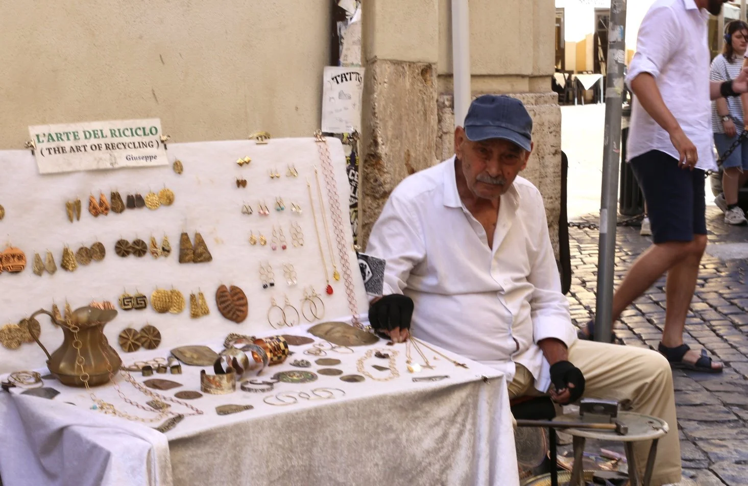 An elderly man sitting beside a table displays jewelry and accessories for sale, with a sign reading 'L'ARTE DEL RICICLO (THE ART OF RECYCLING) Giuseppe'.