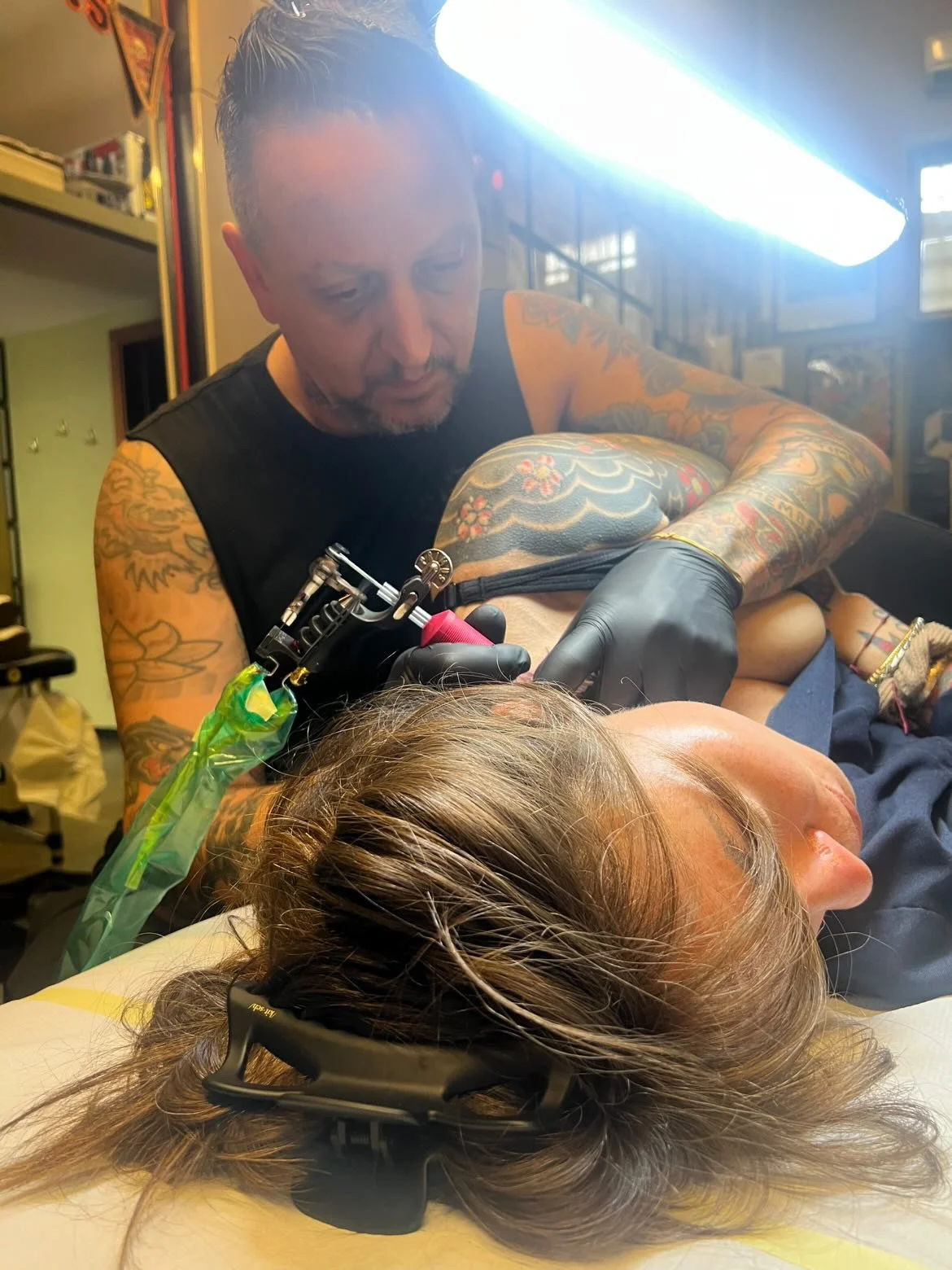 A tattooed man giving a tattoo to a woman lying on her side, with her head resting on a pillow. The woman has long hair and is wearing headphones. The tattoo artist is wearing black gloves and is focused on his work.