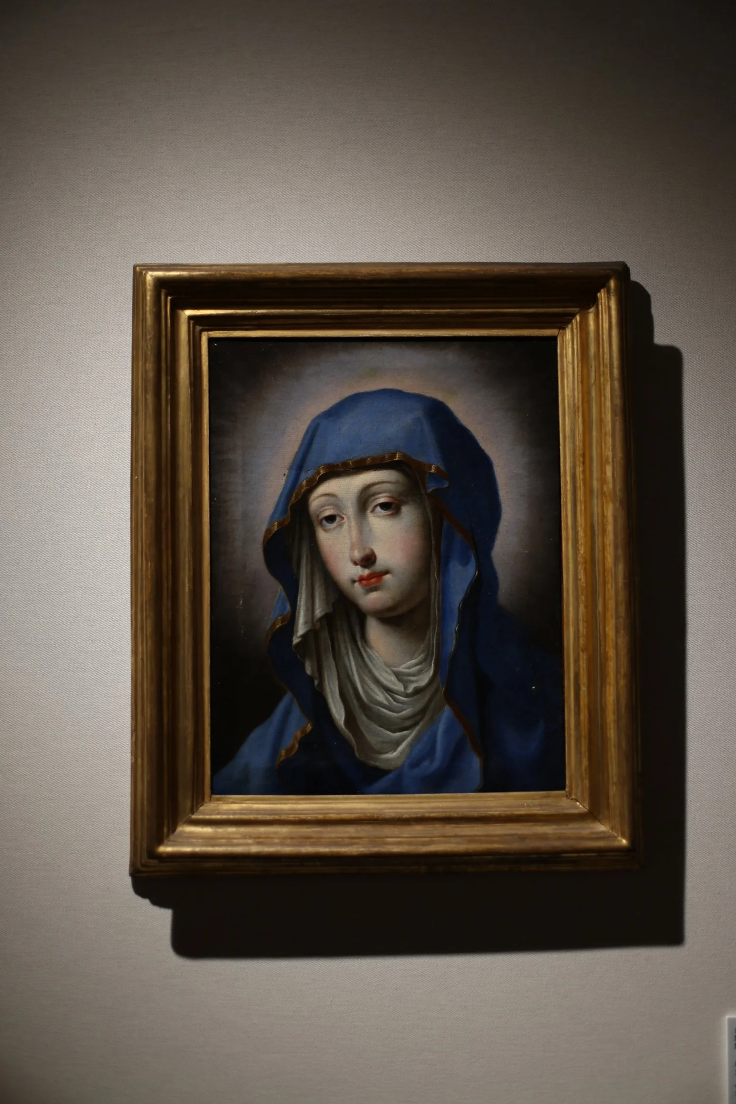 Painting of the Virgin Mary wearing a blue and white head covering, displayed in a gold frame on a gallery wall.