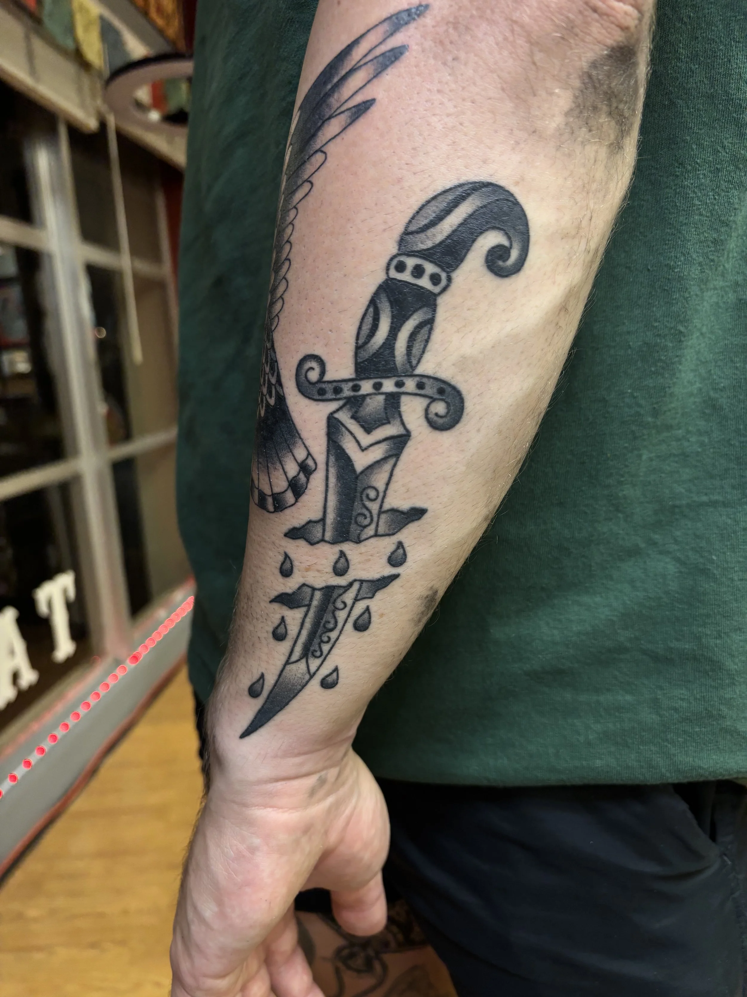 Tattoo of a sword with a decorated hilt and a curved blade, dripping with ink, on a person's forearm.