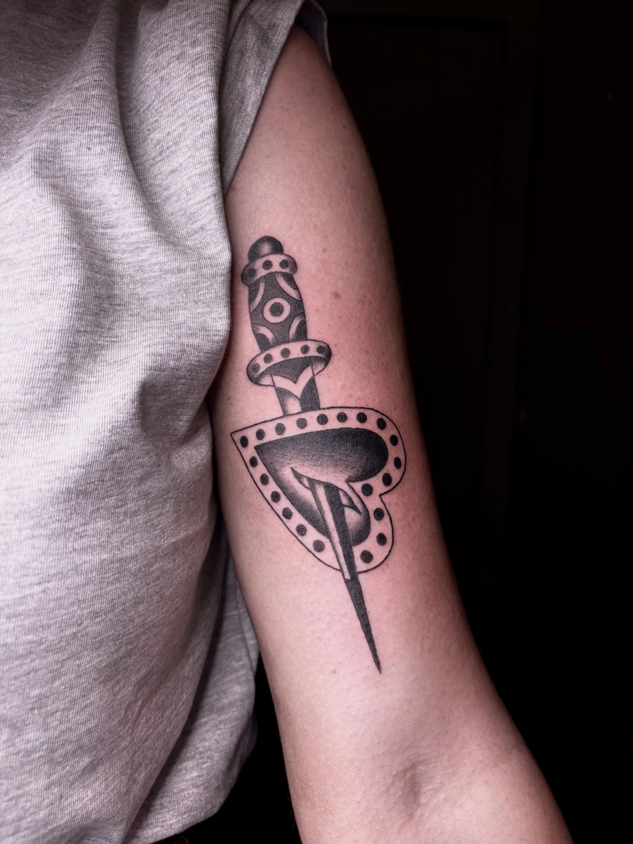 Tattoo of a dagger piercing through a heart-shaped shield on a person's upper arm.
