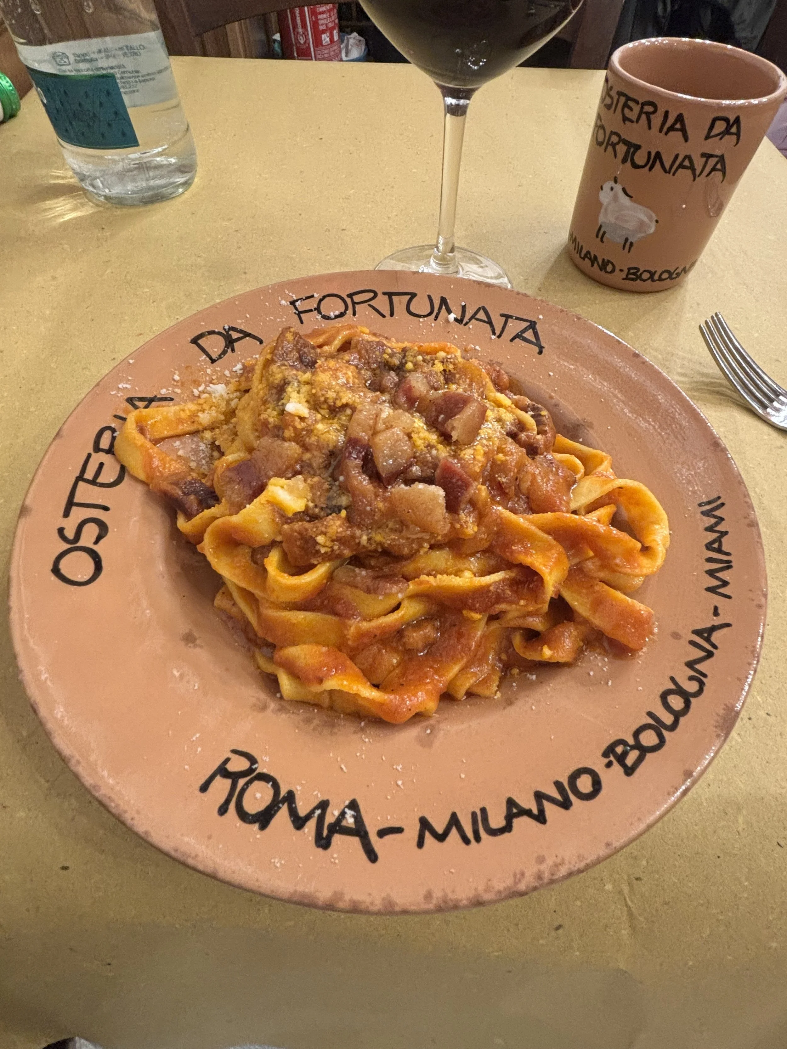 Plate of pasta with tomato sauce and bits of meat, served on a decorative pink plate with black writing around the edge. In the background, there is a glass of red wine, a bottle of water, a pink mug with a goat illustration and Italian words, and a 