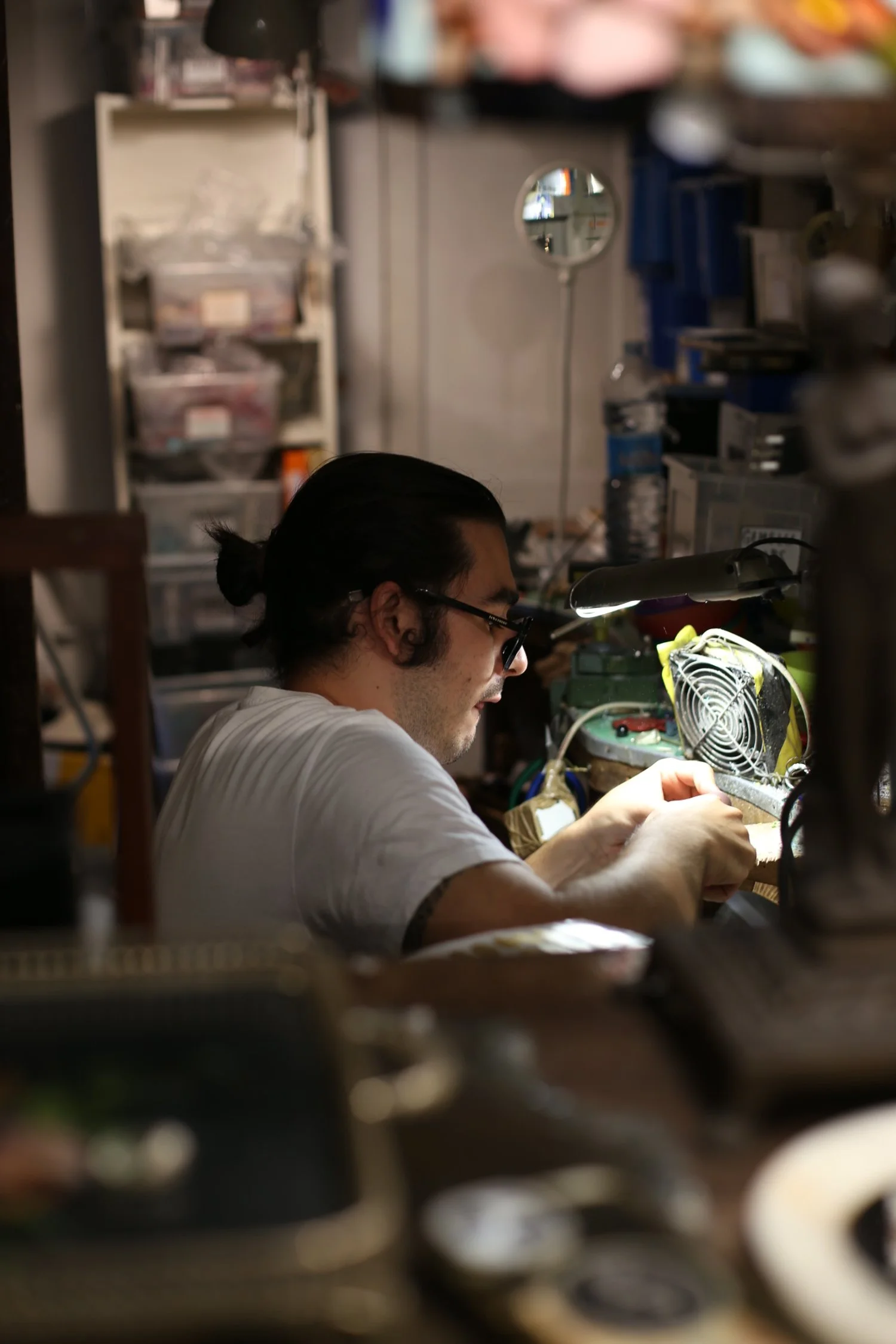 A man with long dark hair tied back, wearing glasses and a white t-shirt, working on an electronic project in a cluttered workshop with various tools and supplies.