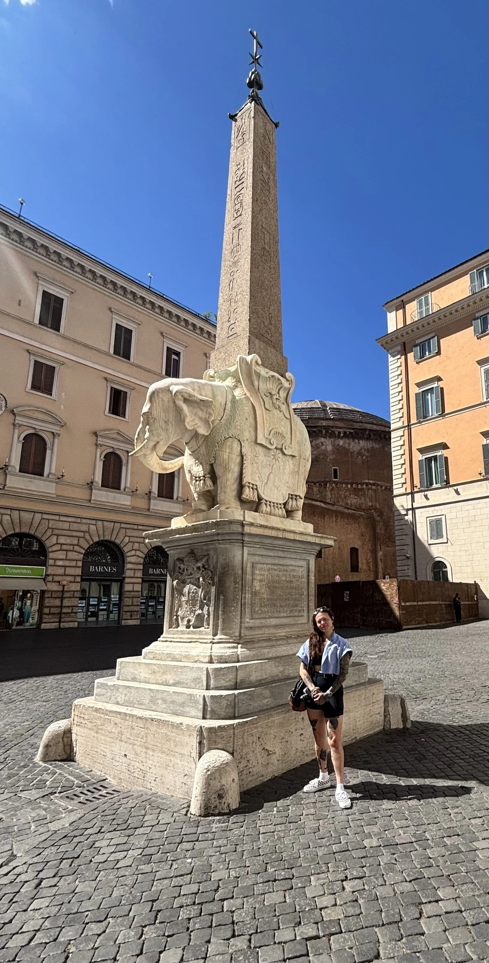 A woman standing in front of an ancient stone sculpture of an elephant on a city square, with an obelisk monument behind her, surrounded by buildings under a clear blue sky.