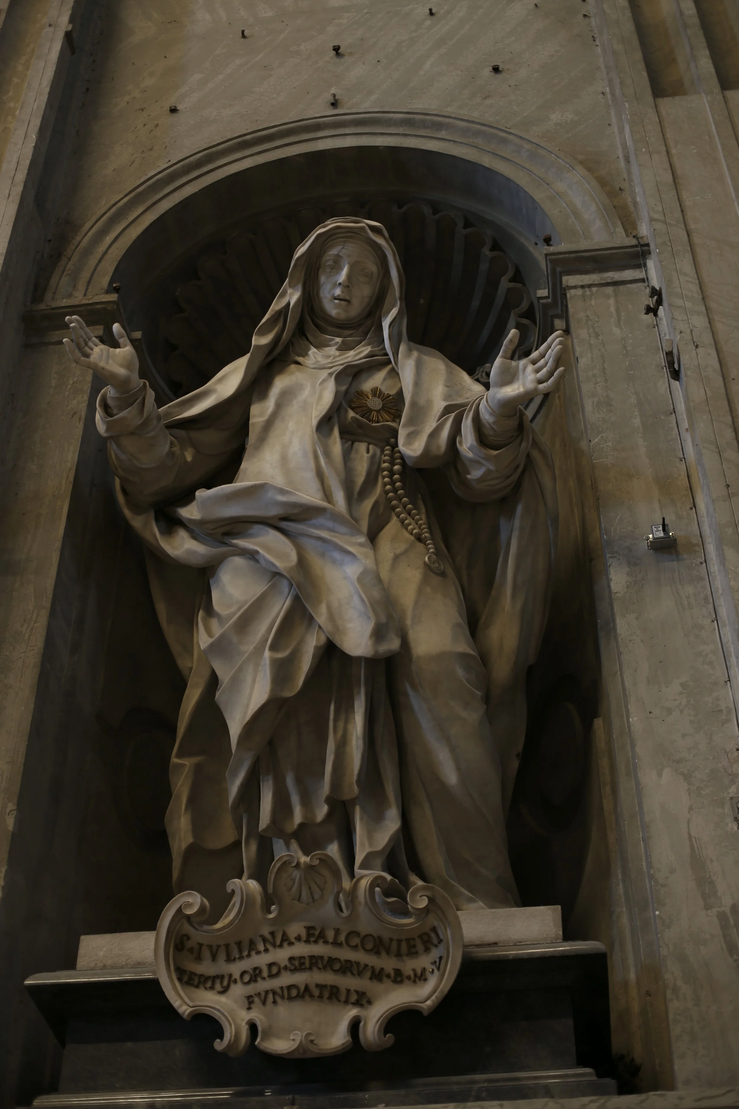 A religious statue of the Virgin Mary with open arms, located inside a cathedral.