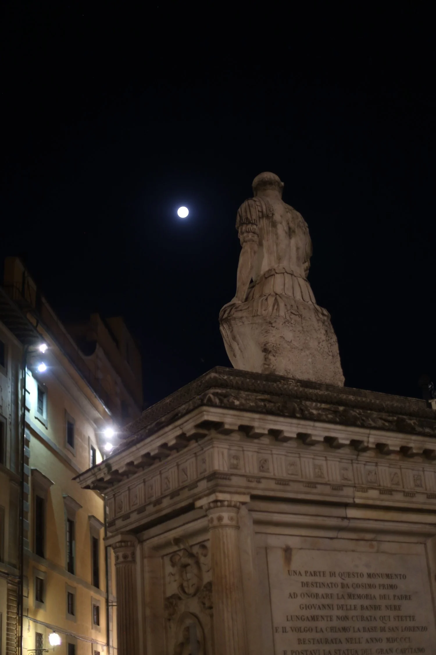 Night view of a historic monument with a statue on top and a plaque at the base, illuminated by nearby lights, with the moon visible in the dark sky.