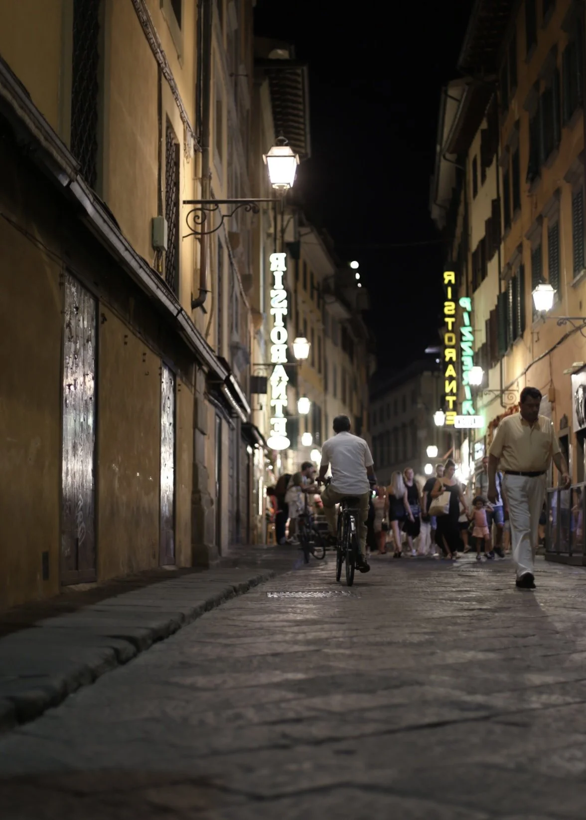 Nighttime street scene with people walking and a man riding a bicycle, illuminated signs on buildings reading 'RESTAURANT' and 'NIGHT' in Italian, old-style street lamps, and cobblestone pavement.