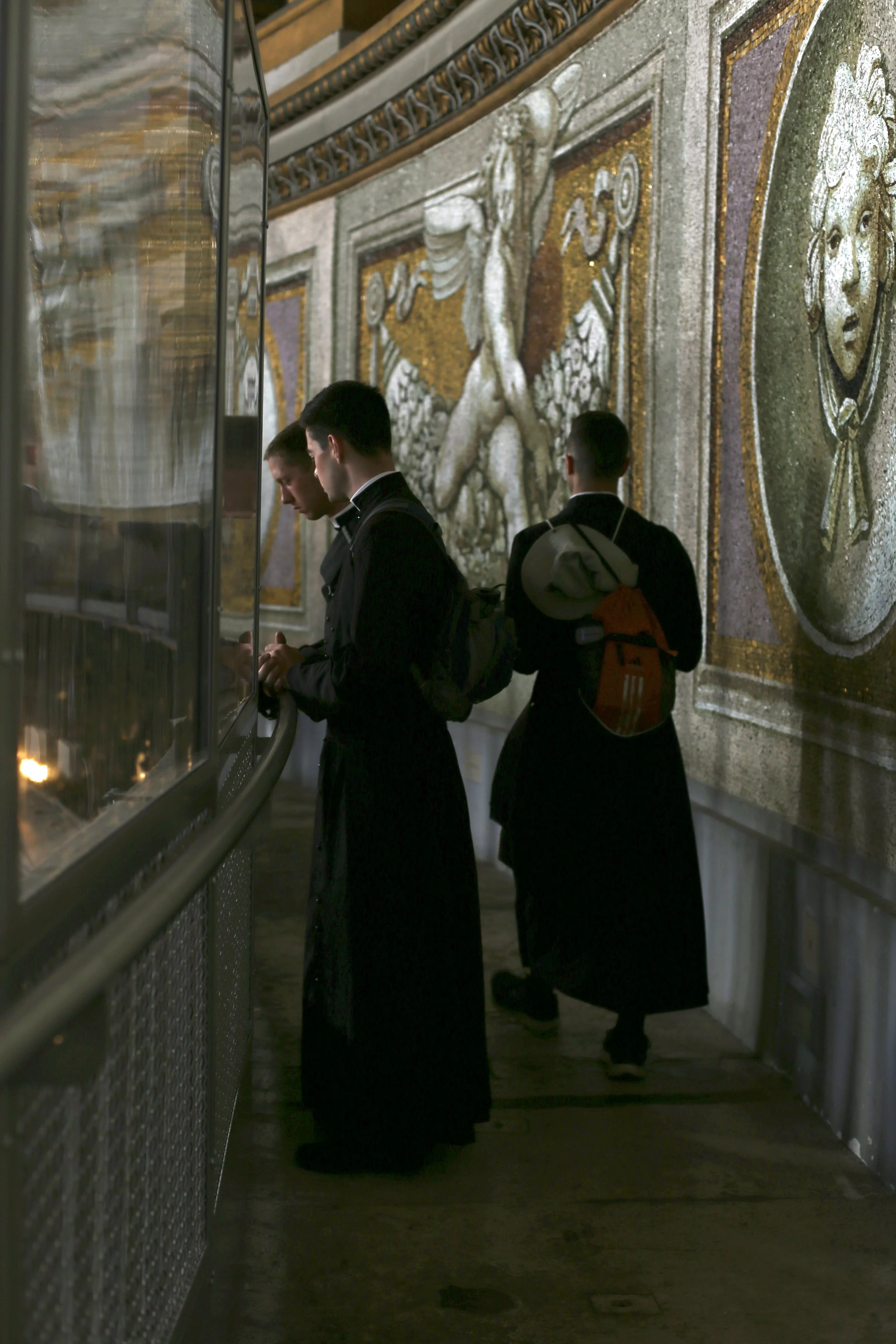 Two men dressed as priests standing in front of a glass display case, with an ornate mosaic wall featuring classical figures and faces in the background.