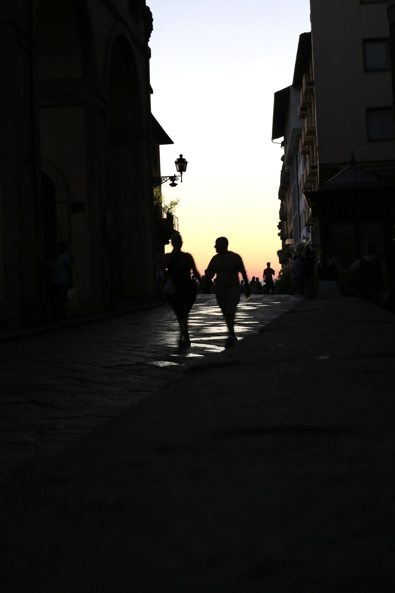 Silhouetted couple holding hands walking down a cobblestone street at sunset, with other pedestrians and buildings lining the street.