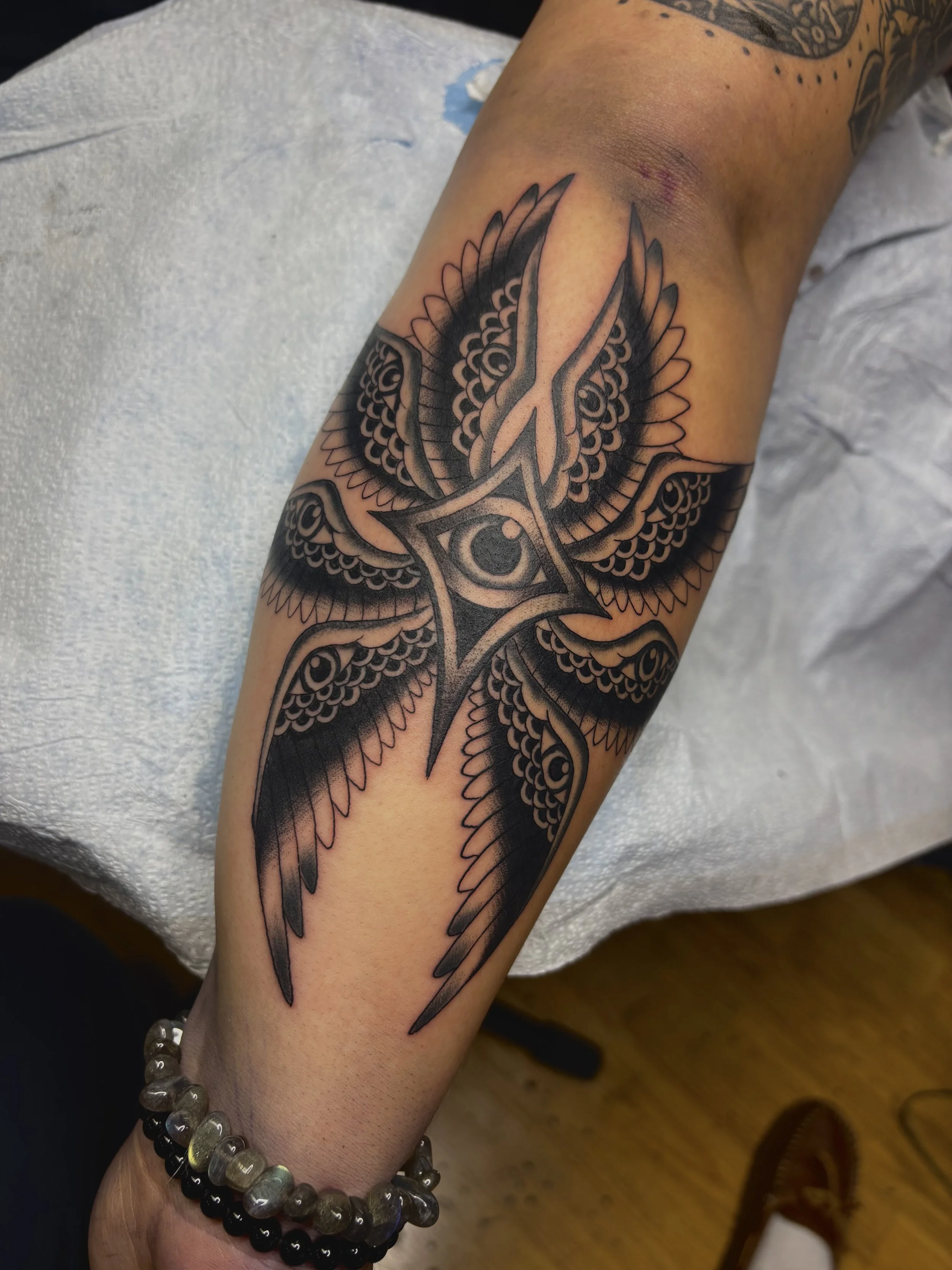Black ink tattoo of a stylized bird with a central eye, surrounded by intricate feather patterns on a person's forearm.