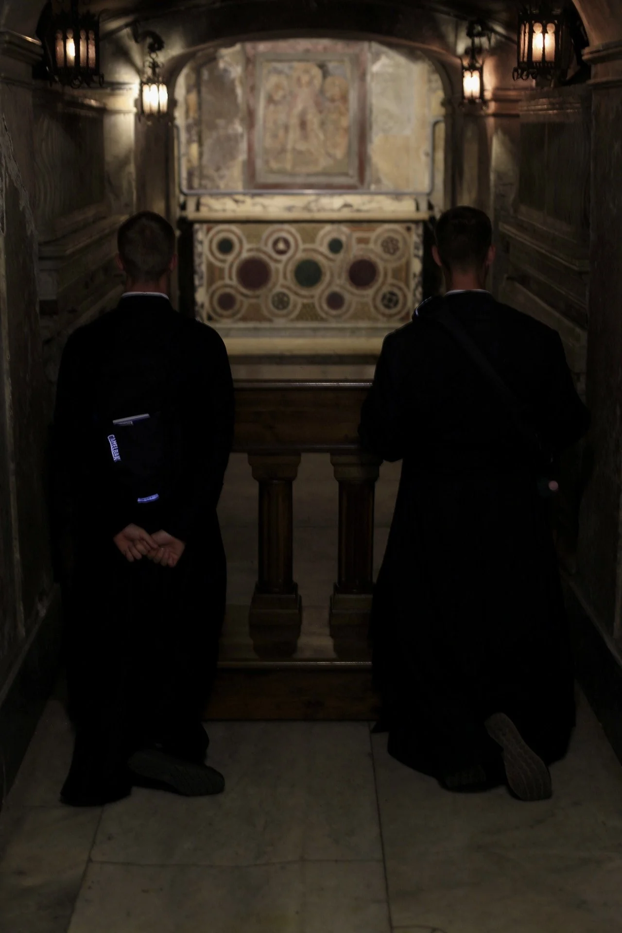 Two people kneeling in prayer inside a church with ornate walls, artwork, and lit wall sconces.