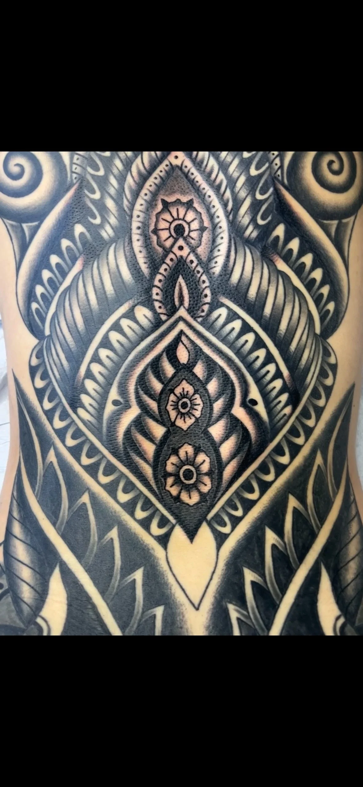 Close-up of an intricate black and grey tribal-style tattoo on a person's chest and abdomen, featuring geometric patterns, floral motifs, and detailed shading.