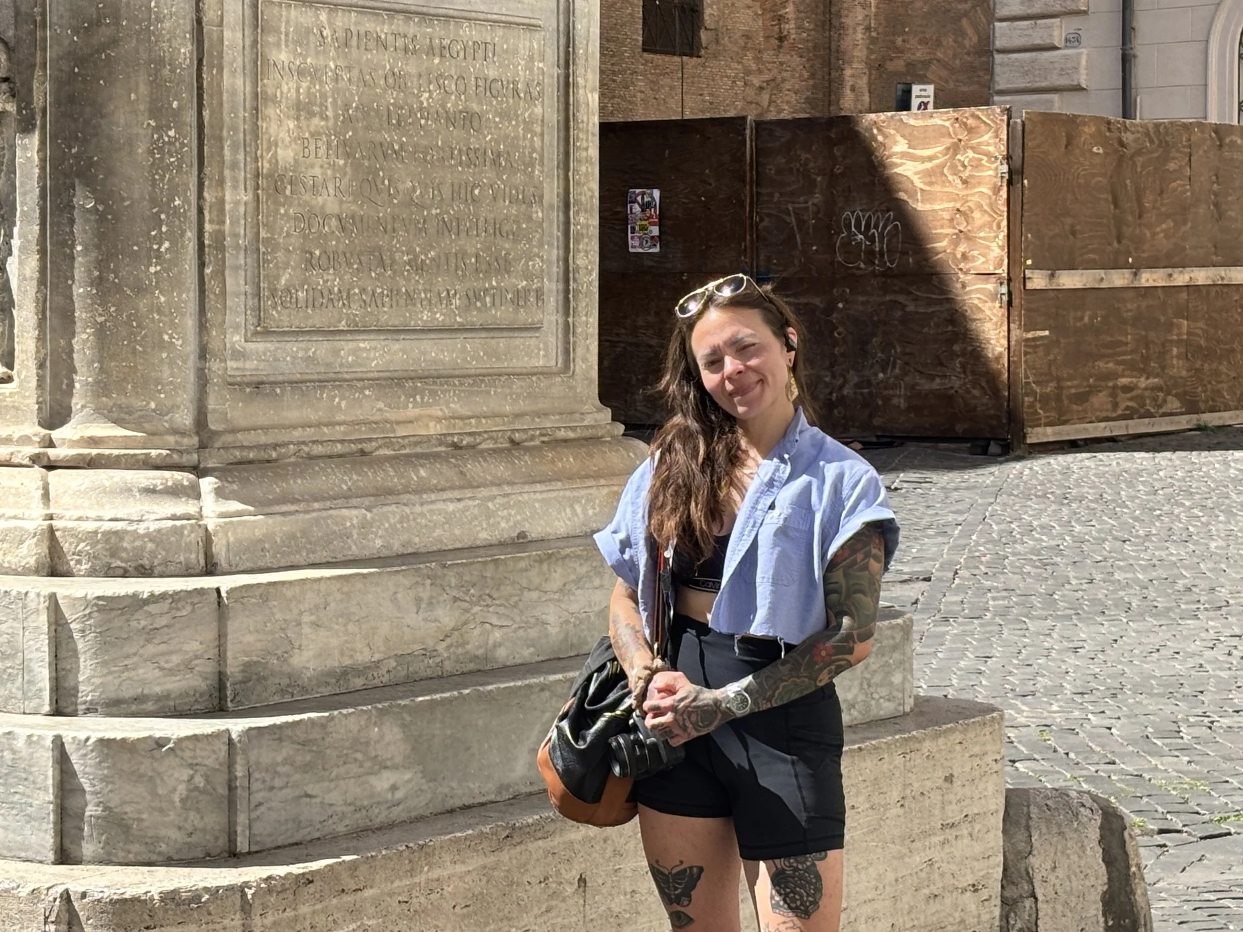 A woman with long brown hair, tattoos on her arms and legs, standing outdoors next to an old stone monument with engraved text. She is wearing sunglasses on her head, a black top, black shorts, and a blue shirt draped over her shoulders, holding a ca