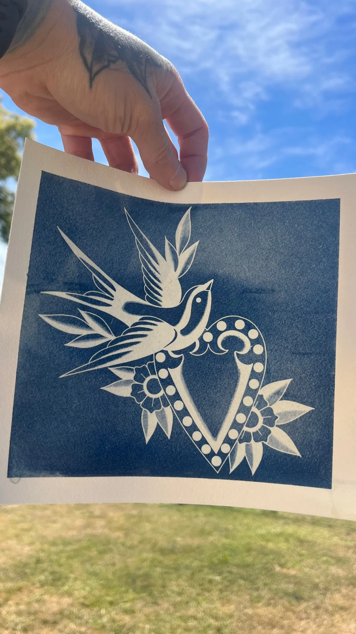 Close-up of a hand holding a paper with a blue and white tattoo of a bird and floral design, against a blue sky with some clouds and a grassy field in the background.