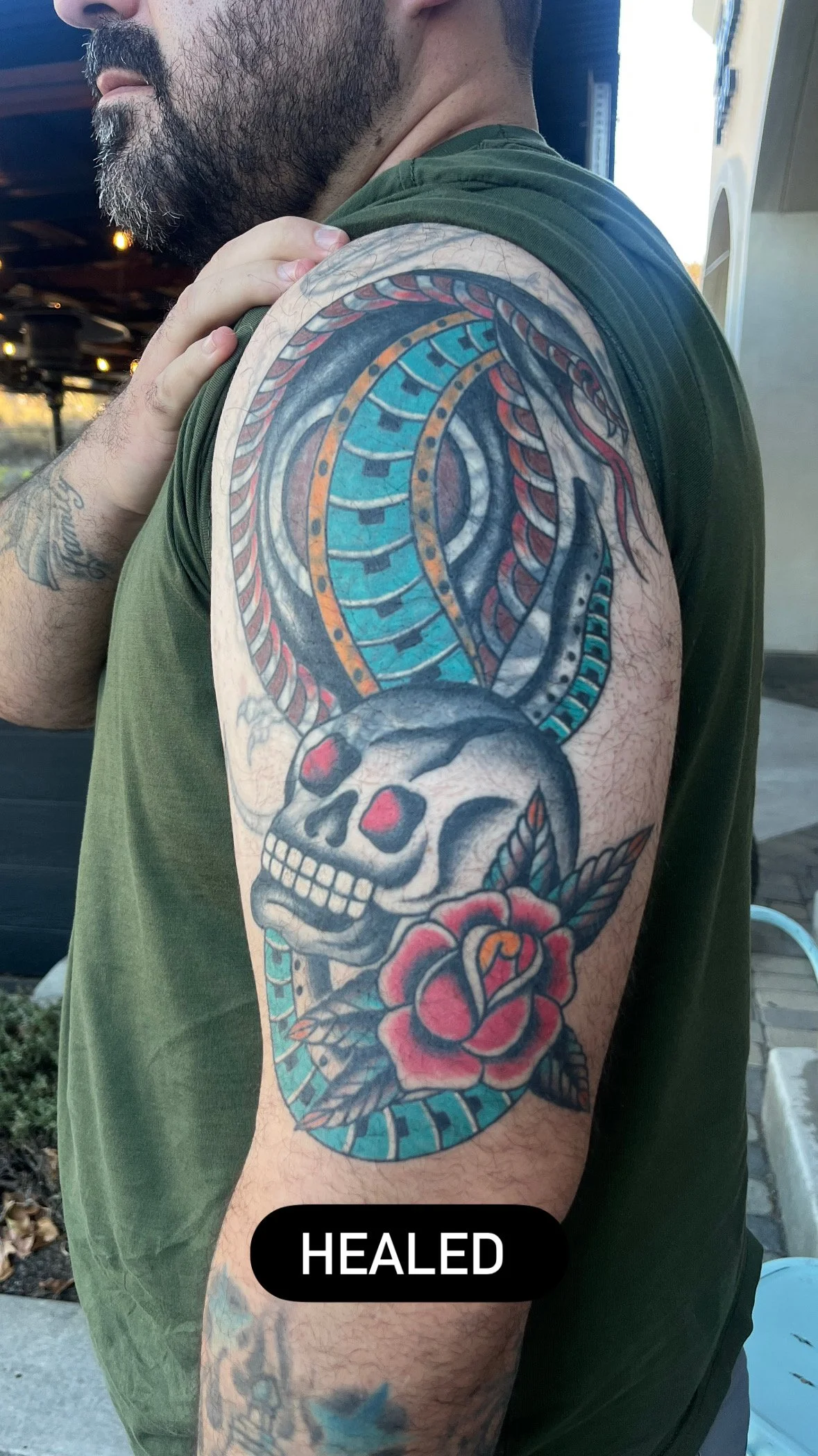 Tattoo of a skull with rose and leaves, and colorful snake coiled around the skull on a man's upper arm.