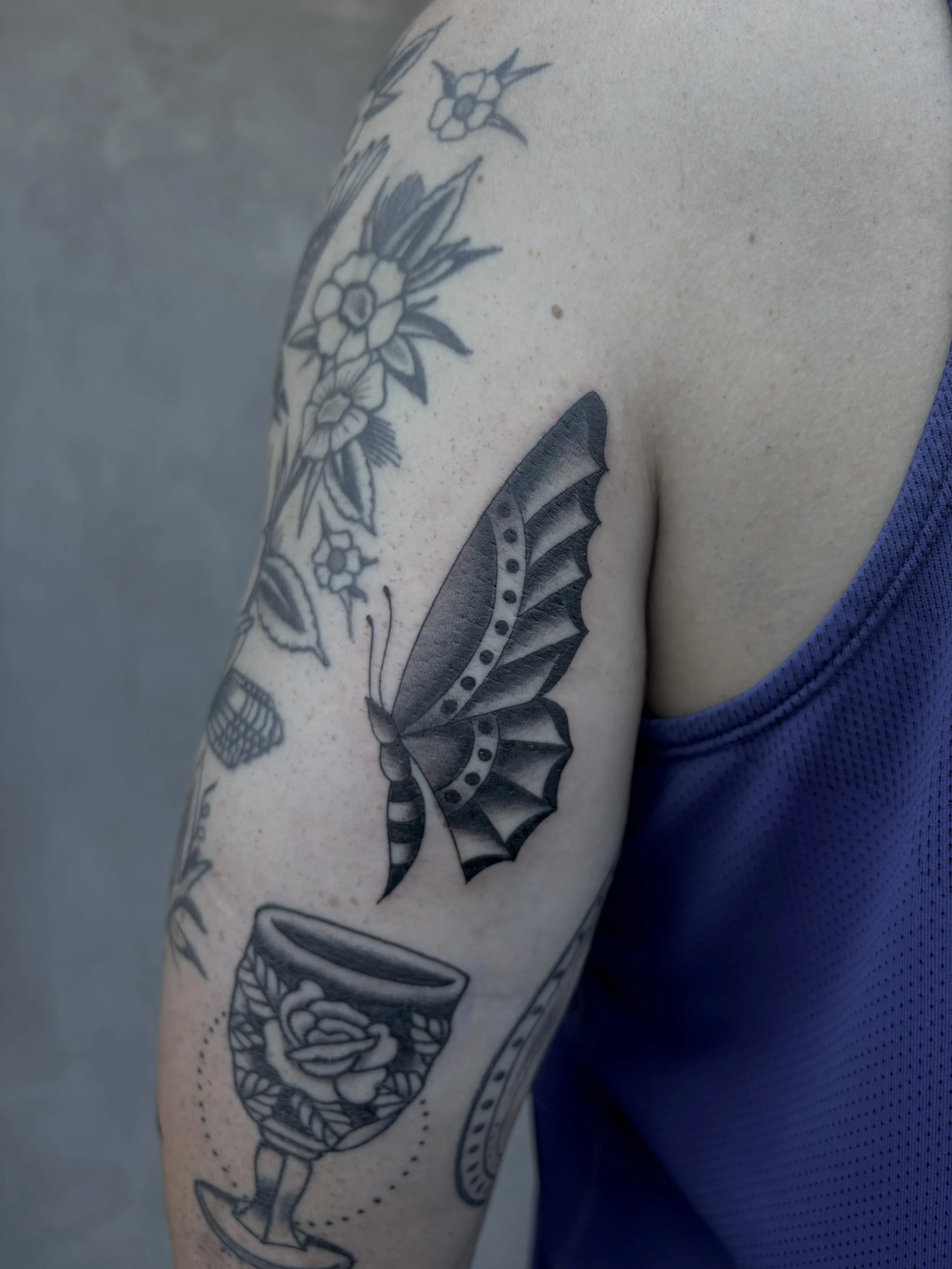 Close-up of a person's upper arm with several black ink tattoos, including a butterfly, a wine glass with a flower motif, and floral designs on the shoulder.